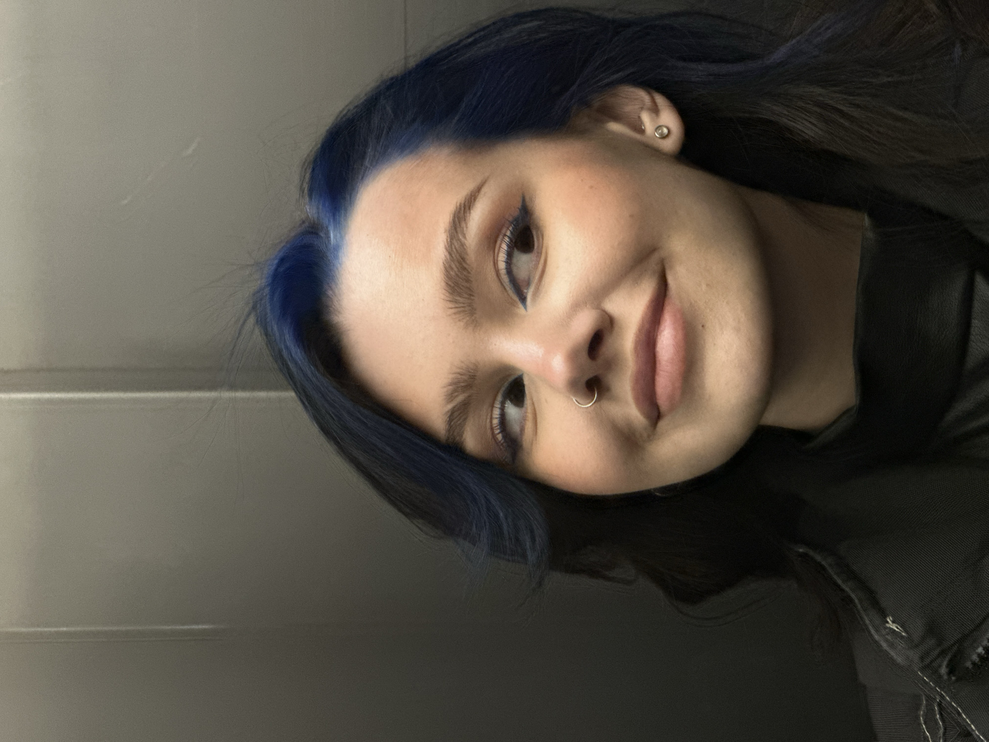 A close-up portrait features a young person, likely female, with distinctive dark blue hair, smiling subtly at the camera. Her head is tilted to the left, showcasing the vibrant blue color of her hair, which appears to be dyed. She has blue eyeliner on her left eye, complementing her hair, and well-groomed eyebrows. A small silver hoop piercing is visible in her right nostril, and a stud earring is present in her left ear. Her lips are naturally colored, forming a gentle, closed-mouth smile. She is wearing a dark, possibly black, garment, with some fabric texture and stitching visible on the right side of the frame. The background is a plain, light grey wall or surface, displaying faint horizontal streaks or light reflections, suggesting an indoor setting. The overall expression is calm and pleasant. There is no visible text. The context indicates the location is Khimki, Russia.