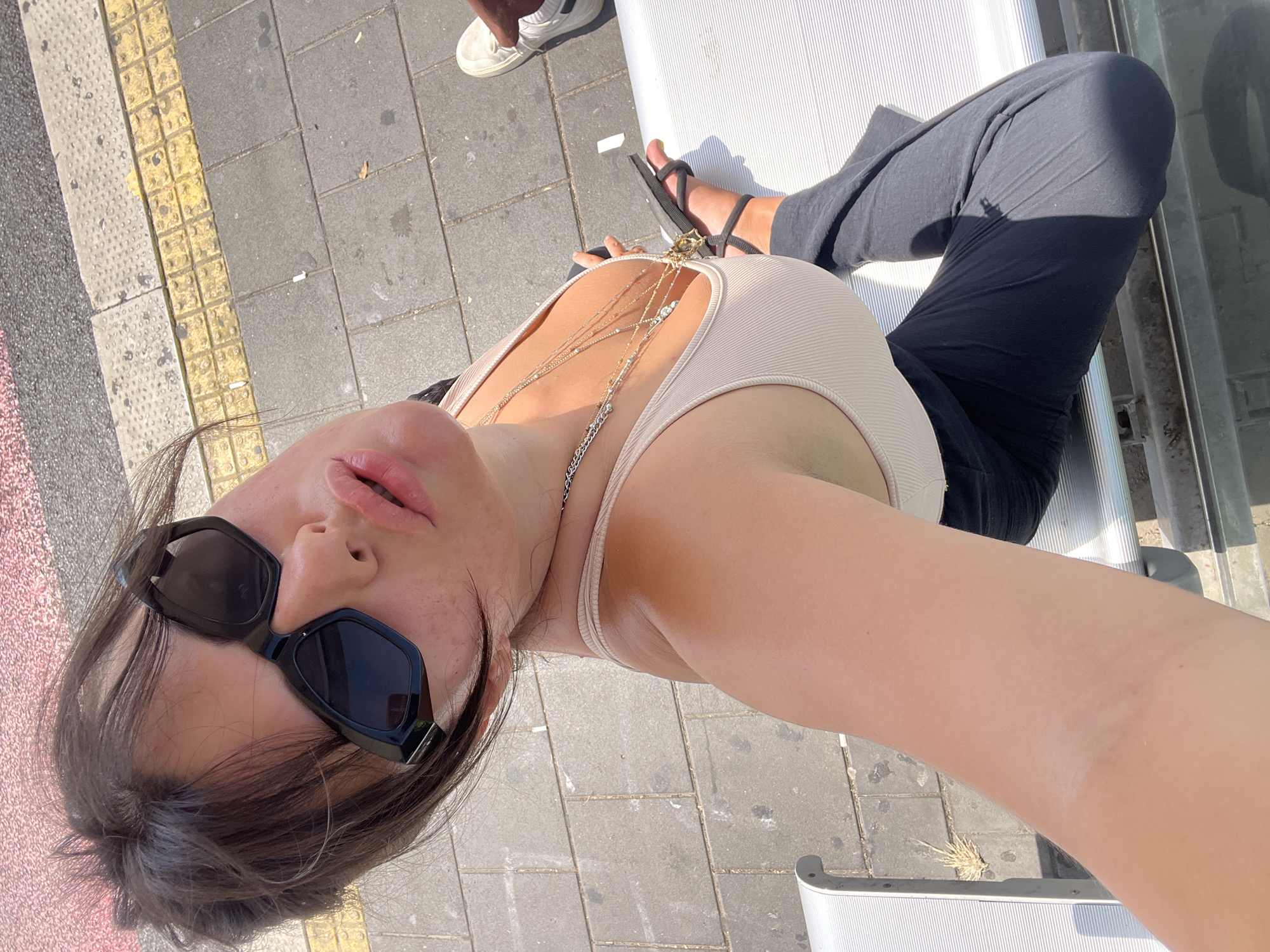 A high-angle, overhead selfie shows a young woman with dark hair, wearing black sunglasses, a light-colored tank top, and dark pants, sitting on a metal bench outdoors. She has multiple necklaces on. Her left arm is extended, holding the camera. The background consists of a paved walkway with yellow tactile paving and red and grey asphalt. A person wearing white sneakers is visible in the upper right corner. The scene is likely a public transportation stop in Kiryat Motzkin, Israel, suggested by the tactile paving common in Israeli public spaces and the architectural style of the bench, which is typical of outdoor public seating in the region.