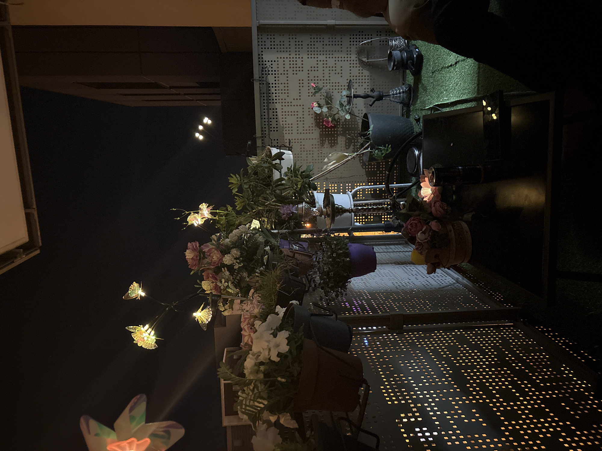 The image shows a nighttime scene in Strasbourg, France. It features a multi-tiered metal structure adorned with potted plants and flowers. The structure is situated on a surface with square perforations that create a dotted pattern due to light passing through. Some of the flowers are illuminated with lights, including some fairy lights shaped like butterflies. Part of a building is visible in the background on the left side of the image. At the top of the image, there is an area covered with artificial turf and what appears to be a surveillance camera.