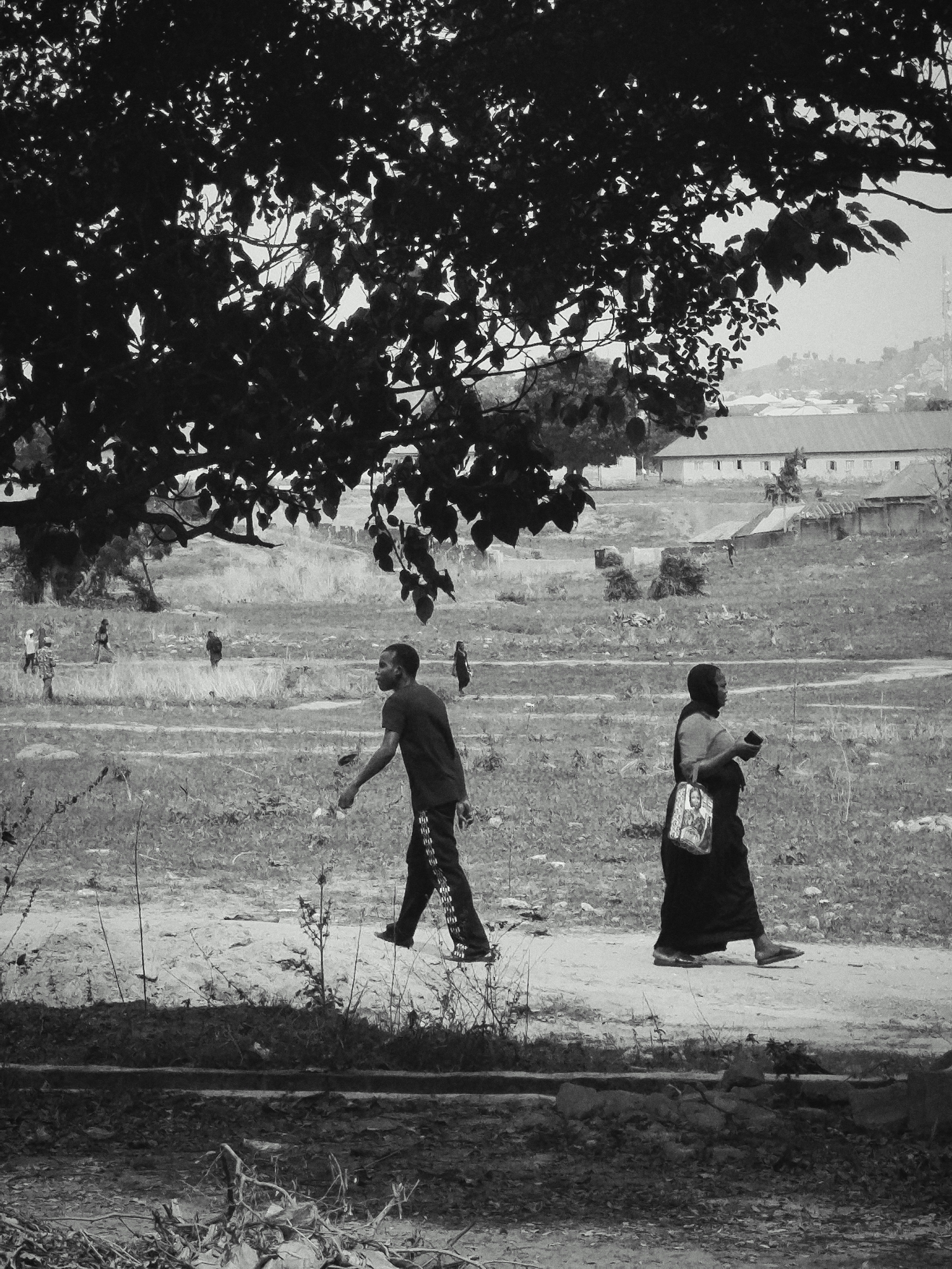 A black and white photograph depicts an open, expansive outdoor scene in Keffi, Nigeria, featuring people walking through a grassy field under a large, leafy tree. The upper left portion of the image is dominated by the dense foliage of a tree, its dark leaves creating a natural frame.
In the foreground, two main figures are walking from left to right on a dirt path that cuts across the landscape. The figure on the left is a young man wearing a dark t-shirt and trousers with a striped pattern. The figure on the right is a woman dressed in a dark garment, carrying a white tote bag that features a prominent religious image, possibly of the Virgin Mary or a saint. She also holds a small dark object in her hand.
In the mid-ground, several other smaller figures are dispersed across the grassy field, suggesting general movement or activity. The background reveals a cluster of buildings with light walls and dark roofs, situated on a gentle hillside, hinting at a village or semi-urban settlement beyond the open field. The sky appears bright but hazy, creating diffused lighting conditions. The overall impression is one of daily life unfolding in a natural, somewhat undeveloped setting. The photograph has a slightly grainy texture. No readable text is discernible, apart from the iconography on the woman's bag.