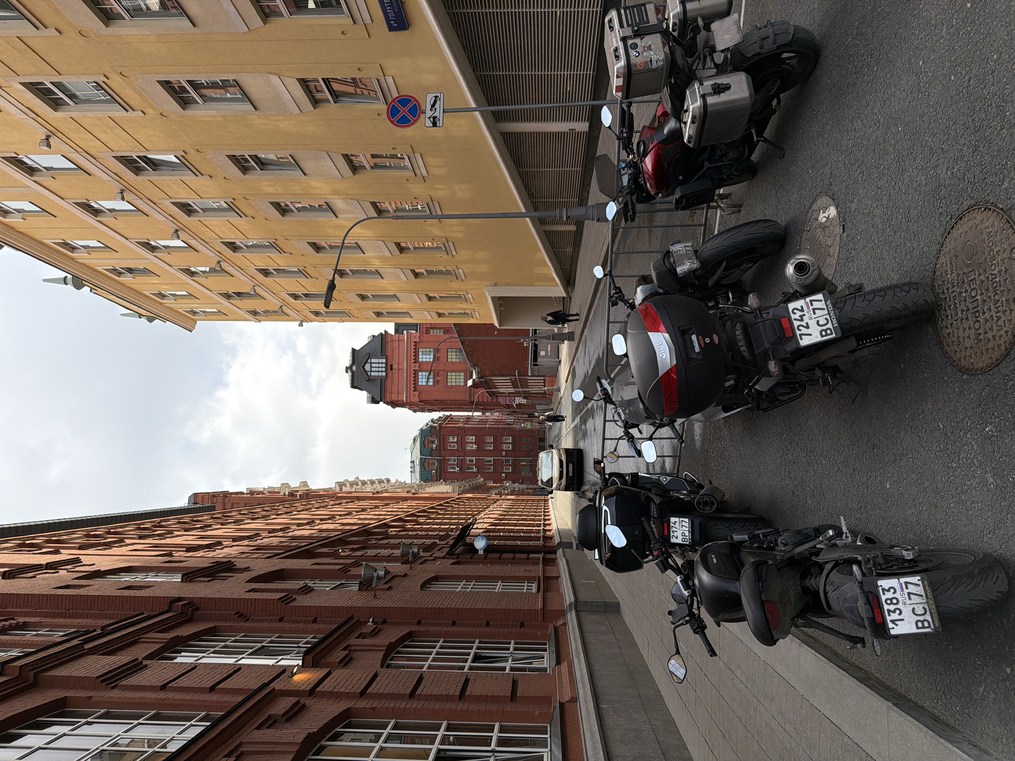 Here's a detailed description of the image:
**Scene Overview:**
The image presents a street scene, seemingly in the city of Moscow, Russia. The buildings lining the street are distinct with their architectural styles. The buildings appear to be historic structures with various architectural details. The street is lined with parked motorcycles, and a car can be seen in the distance.
**Specific Elements:**
1. **Buildings:**
* On the left is a tall brick building with multiple windows arranged in a regular pattern. The facade is ornate with various design elements.
* On the right is a building with a lighter-colored facade, also with numerous windows.
* In the distance, there is a red building.
2. **Vehicles:**
* Several motorcycles are parked on the side of the street.
* License plates are visible on the motorcycles. The license plate numbers visible include 2174 BP 77, 1383 BC 77, and 7242 BC 77.
* A car is seen further down the street between the buildings.
3. **Street and Sidewalk:**
* The street appears to be paved.
* A sidewalk borders the brick building on the left.
4. **Street Furniture/Signage:**
* A street sign is visible with a "no parking" symbol on a pole extending from the building on the right.
* Manhole covers are visible in the street.
5. **General Appearance:**
* The overall lighting suggests it is daytime.
**Location and Context:**
The license plates are Russian, indicating the scene is in Russia. One of the street signs mentions "4 ГОЛУТВ ПΕΡΕУ," which indicates that the street is the 4th Golutvinsky Lane in Moscow, Russia.