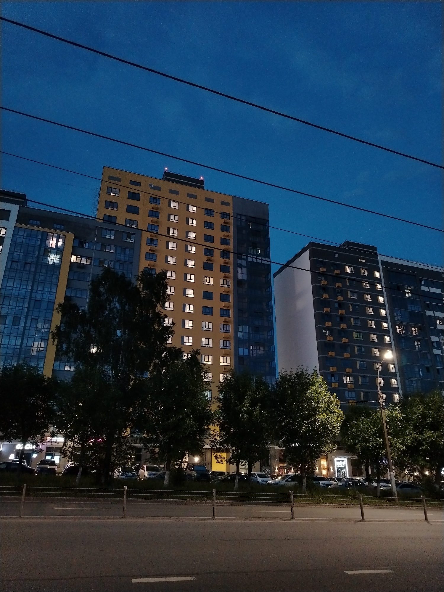 The image displays an urban night scene, likely at late dusk or early evening, characterized by a deep, dark blue sky. Three multi-story buildings form the main subjects in the background. The most prominent is a tall, yellow/ochre-colored building with numerous windows, many of which are illuminated, suggesting occupied residences or offices. To its left, another building with a darker, reflective facade and lit windows is partially visible. On the right, a third tall building features a darker exterior with a distinct vertical white section, also showing many illuminated windows.
In the mid-ground, a line of dense, dark green trees partially obscures the lower parts of the buildings and a row of parked cars. A bright street light is visible on the right, casting a localized glow. Stretching across the top portion of the frame are two dark power lines or cables.
The foreground consists of an asphalt road with faint white lane markings and a low barrier made of posts and rails, separating the road from a grassy median. Behind the trees, glimpses of storefronts with interior lights are visible, indicating ground-floor businesses. No explicit human activity is captured, but the illuminated windows and parked cars imply a living, active urban environment. The general atmosphere is quiet and still, indicative of an evening in a residential or mixed-use area. While specific text on businesses is largely illegible due to distance and lighting, the scene portrays a typical urban setting.
