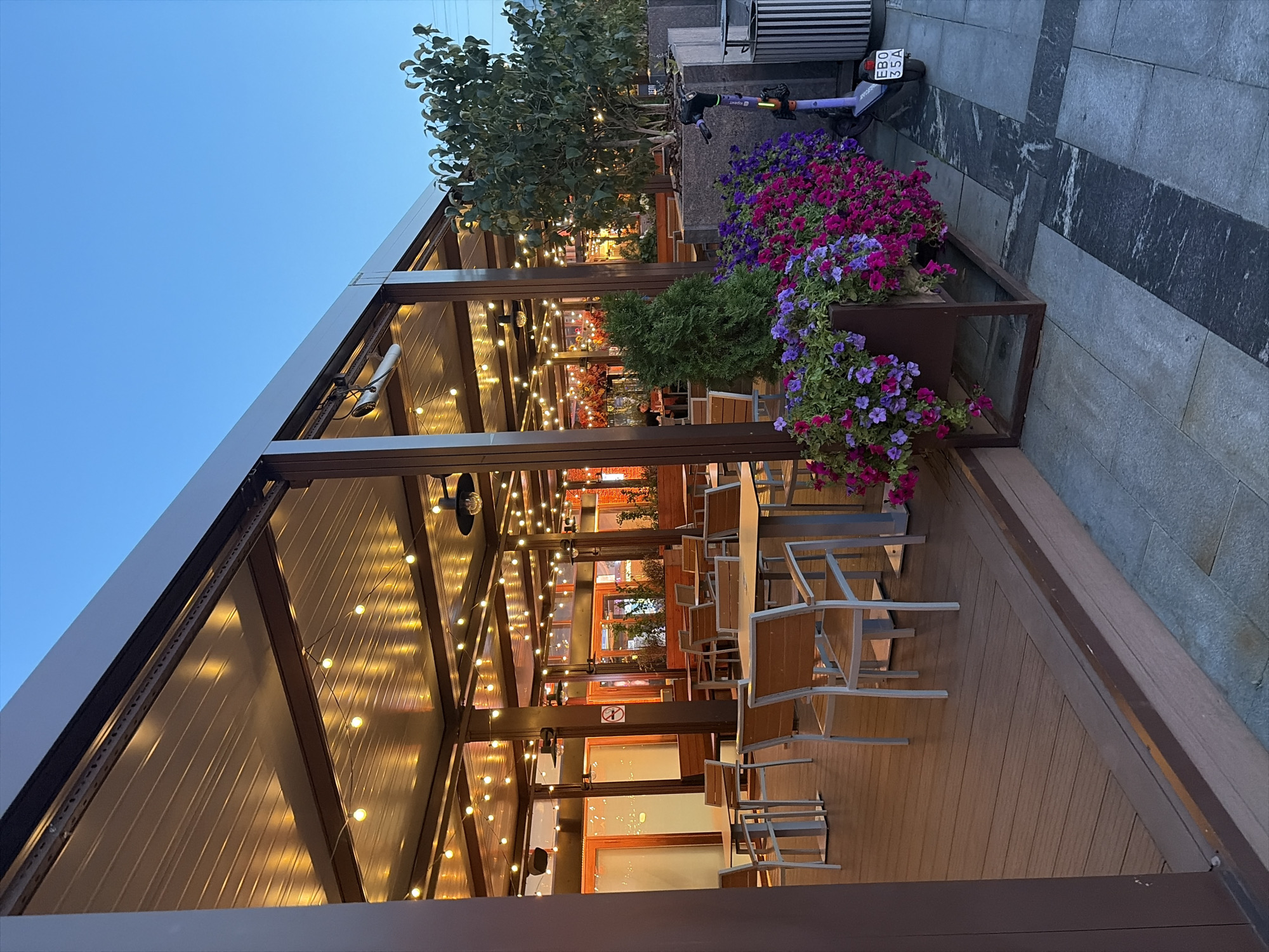 An outdoor dining area with a covered patio and string lights is pictured at dusk in Moscow, Russia. The patio is furnished with wooden tables and chairs, and the floor is made of wooden planks. In the foreground, a planter box overflows with colorful flowers, and a purple electric scooter with a license plate reading "E80 35A" is parked nearby. The background shows the interior of a restaurant with warm lighting and more tables. The sky is a clear twilight blue.