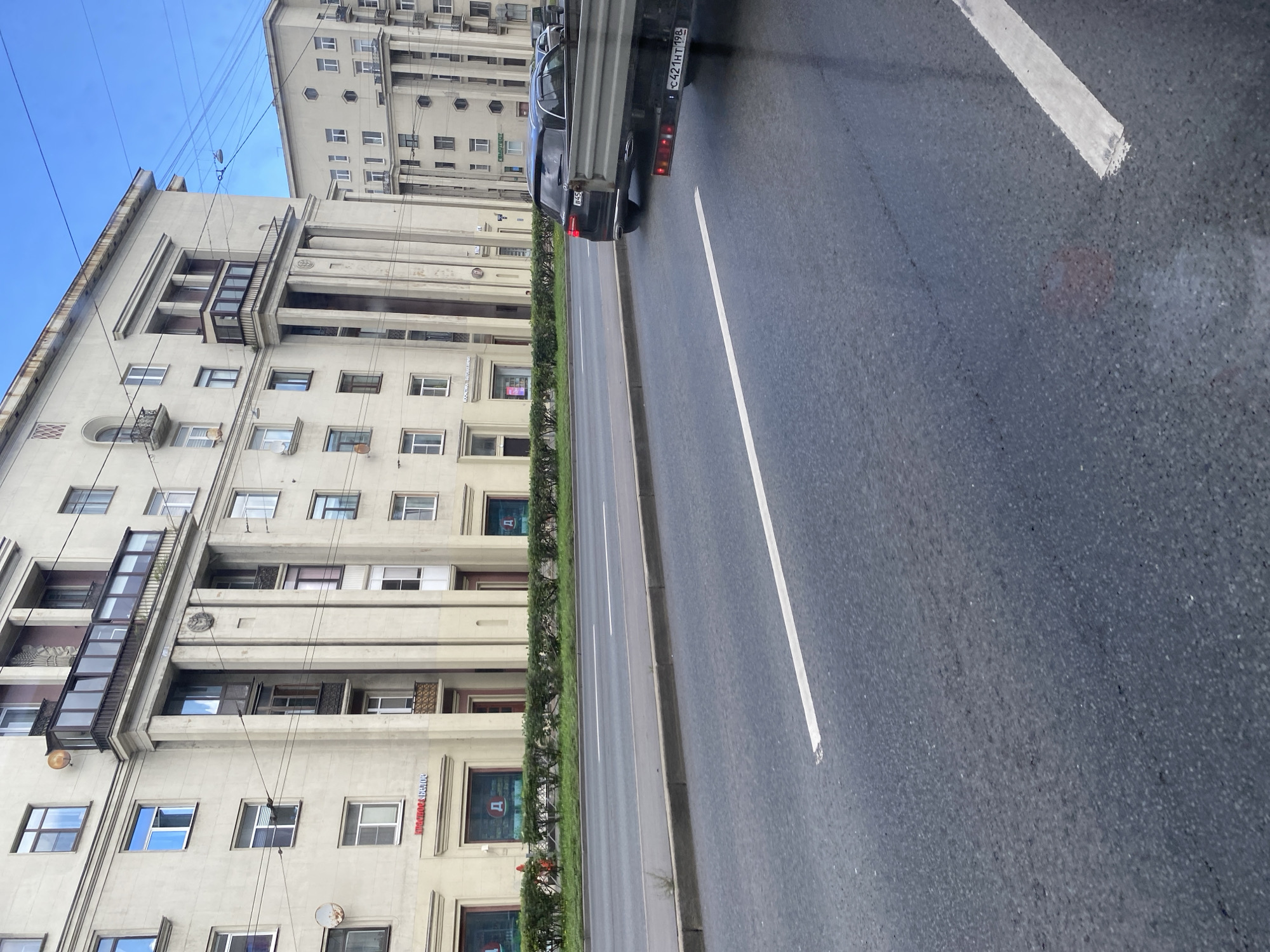 The image depicts a wide, paved street in Saint Petersburg, Russia, with a multi-story, light-colored building lining the left side of the road. The building features numerous windows, balconies with dark railings, and decorative columns. A line of green bushes separates the building from the sidewalk. On the right side of the street, a black SUV with license plate B45 is visible, along with the rear of a truck with license plate C421HT 198. The sky above is overcast with blue and white clouds. The road itself is marked with white lines, indicating lanes and the edge of the roadway.
