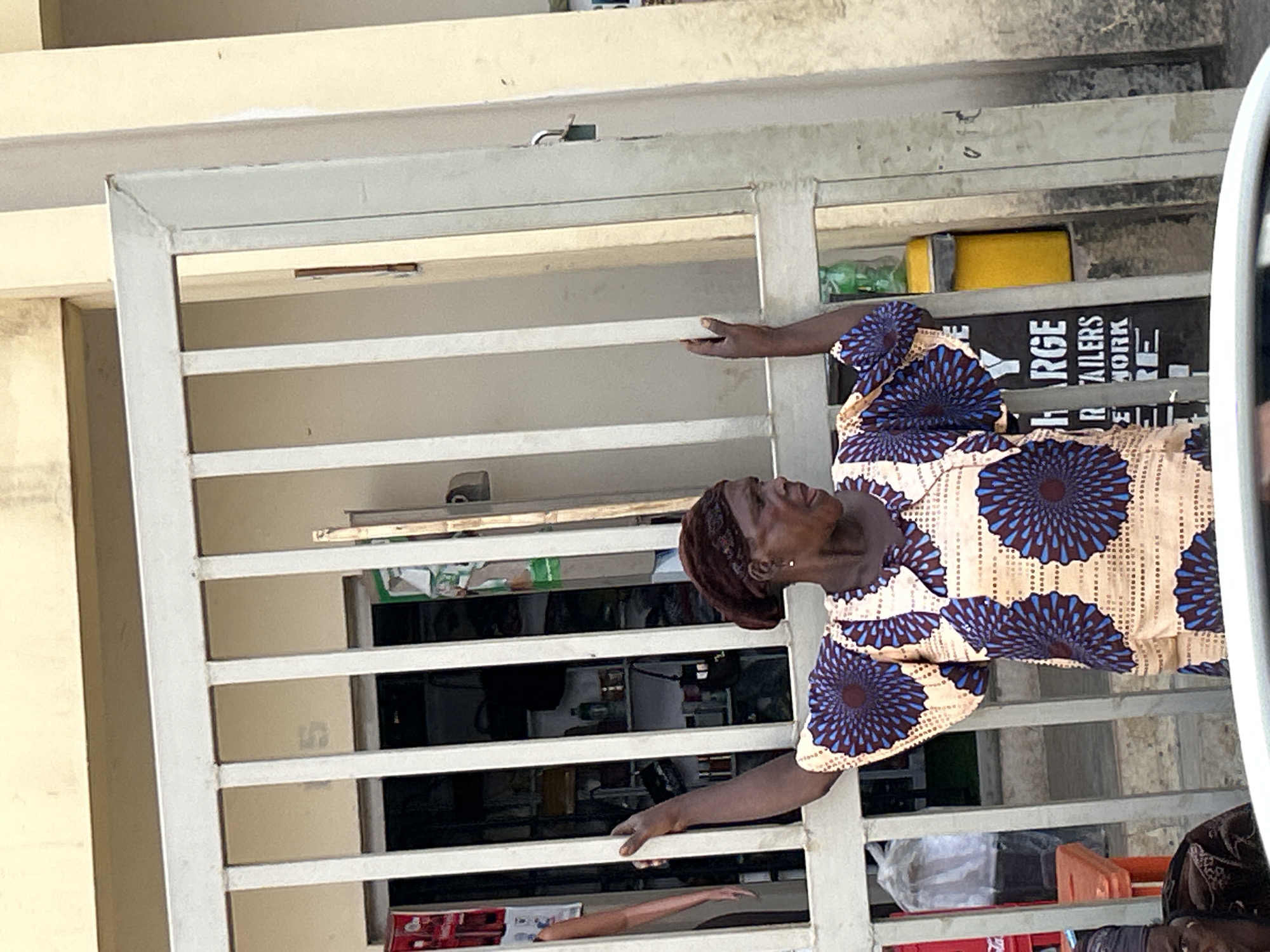 A woman wearing a blue and tan print dress stands behind a white metal gate. Her hands are on the gate, and she is looking to the left. The gate provides a view into the shop behind it. The exact location and date is not provided but the image depicts an environment that resembles a market scene in the city of Jos, Nigeria.