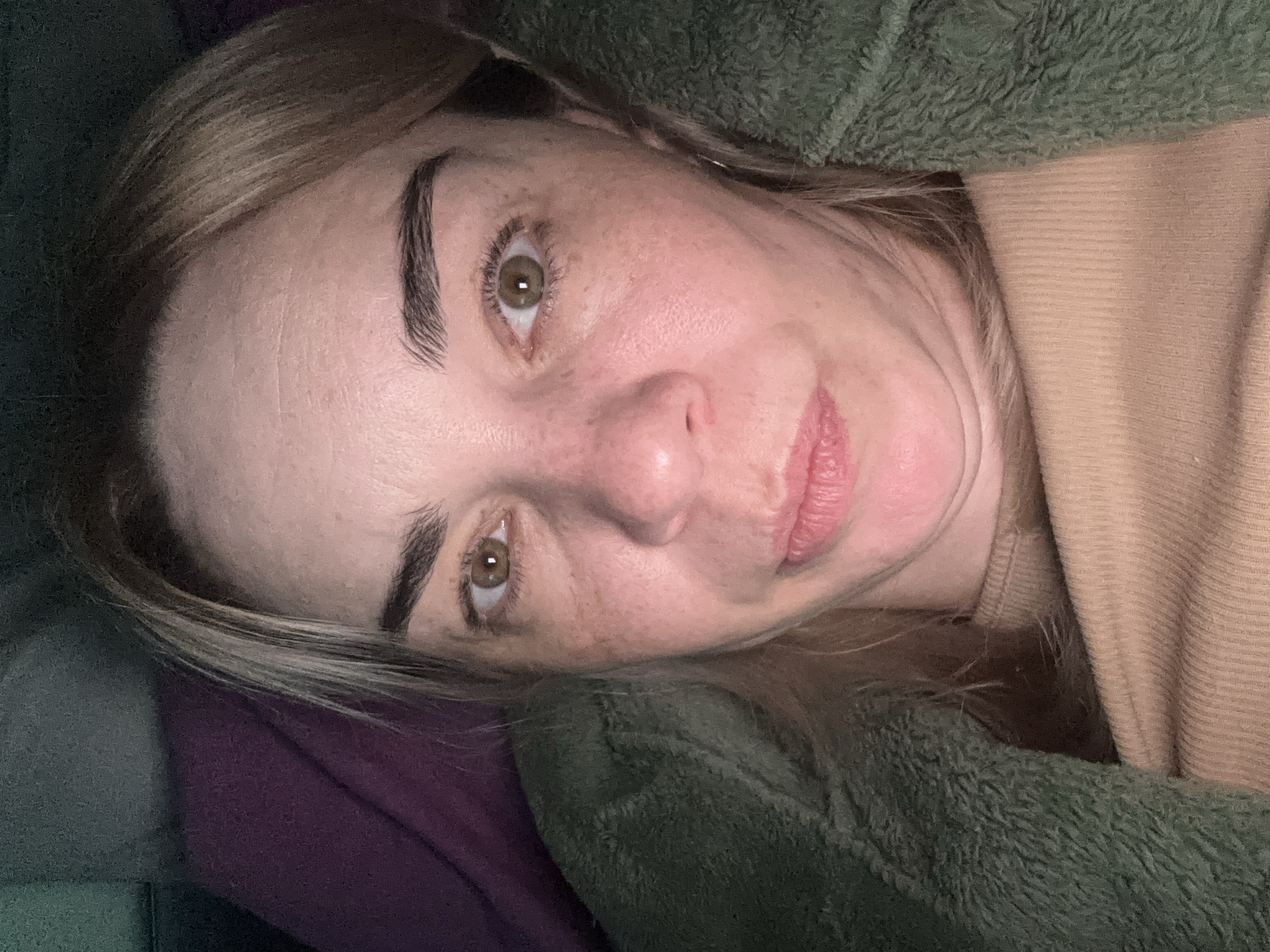 A close-up, slightly rotated portrait shows a woman with fair skin, light brown hair, and freckles. She has arched eyebrows and green eyes. Her lips are pink and slightly parted. She appears to be lying down, with her head resting on a purple pillow or cushion. A soft, green fuzzy material surrounds her face and upper body, likely a blanket or jacket. Part of a beige-colored garment is also visible. The location is unknown, Russia.