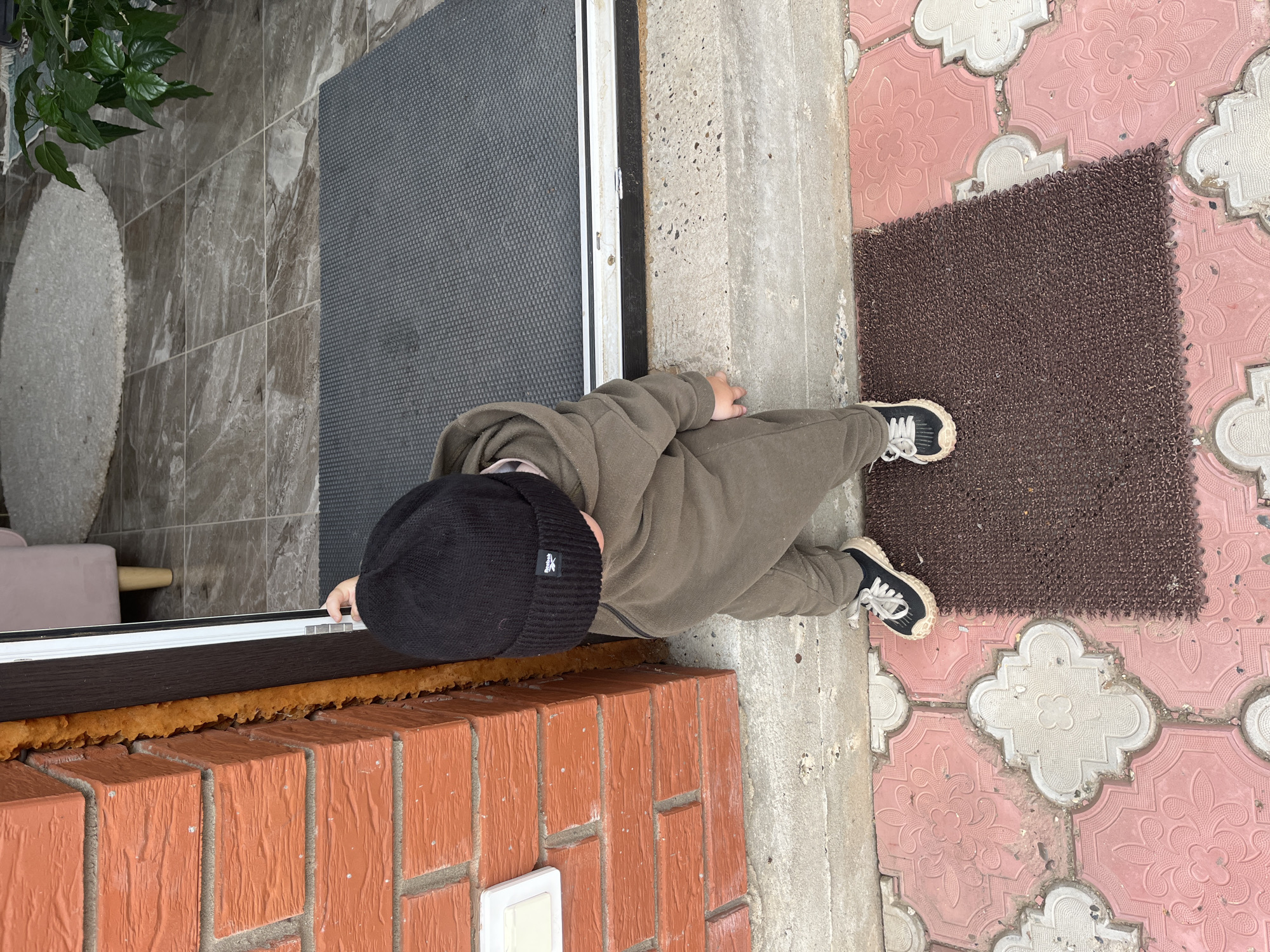A person wearing a black beanie, olive green sweatsuit, and sneakers stands outside a building at what appears to be an entrance. The entrance has a gray door, a brick wall below, and a threshold sealed with yellow foam. The person's hand is on the door frame. A gray mat is inside the entrance. Outside, there is a brown welcome mat on top of pink-patterned paving stones. Green leaves of a plant are visible to the left, next to the interior tiling.
There is no information in the image to confirm the location as Chaikovsky, Russia.