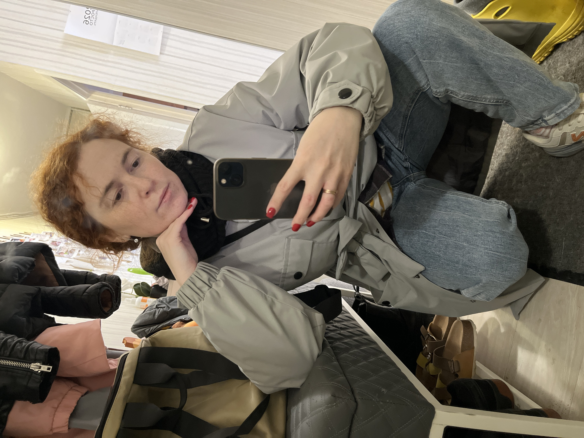 A person is depicted in a selfie pose within a room, presumably indoors. The individual has fair skin, reddish-brown hair, and is wearing a grey jacket and blue jeans. They are holding a dark smartphone, with red nail polish visible on their fingers. A gold ring is worn on one hand. The background contains elements such as clothing items, including jackets and bags, potentially indicating a storage area or closet. A pair of sandals is visible near the lower portion of the frame. Based on the label "ДОСЛОС" which is visible on the sticker it appears that this location could be in Chaikovsky, Russia.