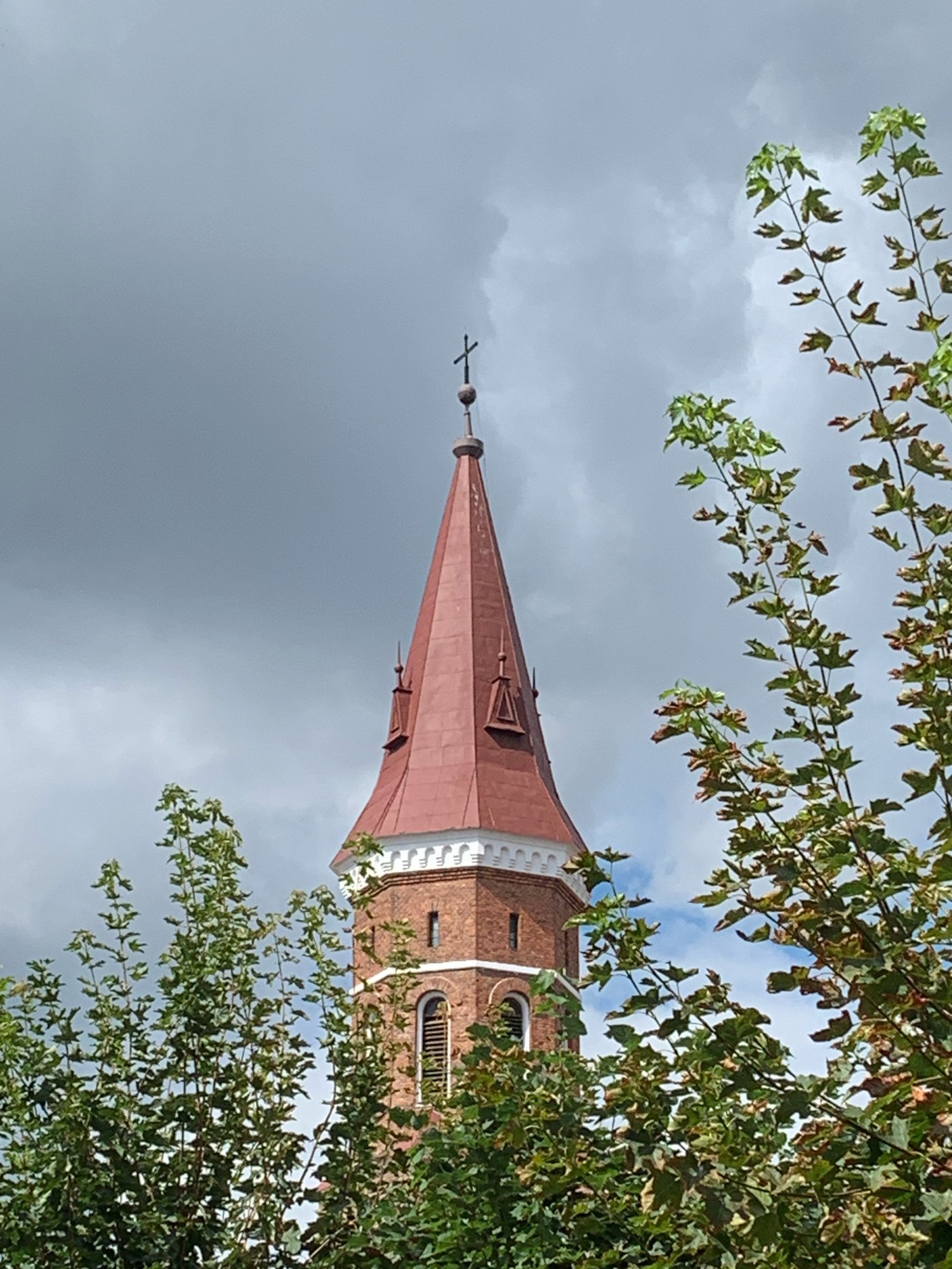 The image captures a close-up view of the upper section of a brick church tower with a red conical roof, topped with a cross. The tower features arched windows with decorative louvers and a white decorative band encircling the base of the conical roof. Lush green foliage from trees partially obscures the lower part of the tower. The sky is overcast with dark, heavy clouds, suggesting an impending storm or recent rain. The scene is likely set in Kolomyia, Ukraine, a city known for its historical churches.