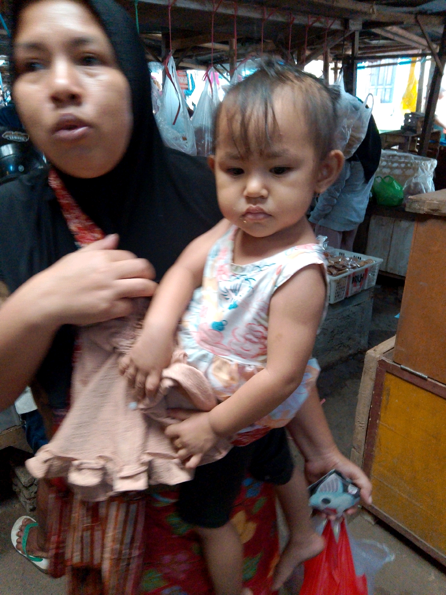 An adult woman wearing a black hijab and a patterned red and white fabric over her shoulder is holding a young child who is facing the camera. The child is wearing a sleeveless, floral-patterned dress and has short, dark hair. The woman's left arm is supporting the child, and her right hand is holding a piece of light-colored fabric with ruffled edges. Both are in what appears to be an outdoor market setting. In the background, there are wooden structures, hanging bags, and various goods displayed in baskets and on shelves. The ground is dirt and debris. The scene is likely in Kediri, Indonesia, given the context provided. The overall lighting suggests it is daytime.