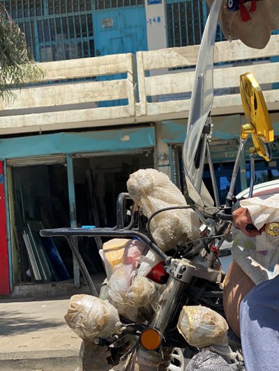 A motorcycle is parked in front of a glass shop in Kandahar, Afghanistan. The front of the motorcycle is heavily wrapped in protective materials like bubble wrap. The shop behind the motorcycle has a blue and white facade with the number "-148" visible above the entrance. Glass panes are stacked inside the shop. The motorcycle's windshield and golden mirrors are noticeable. A hand holding the motorcycle's handle is visible.