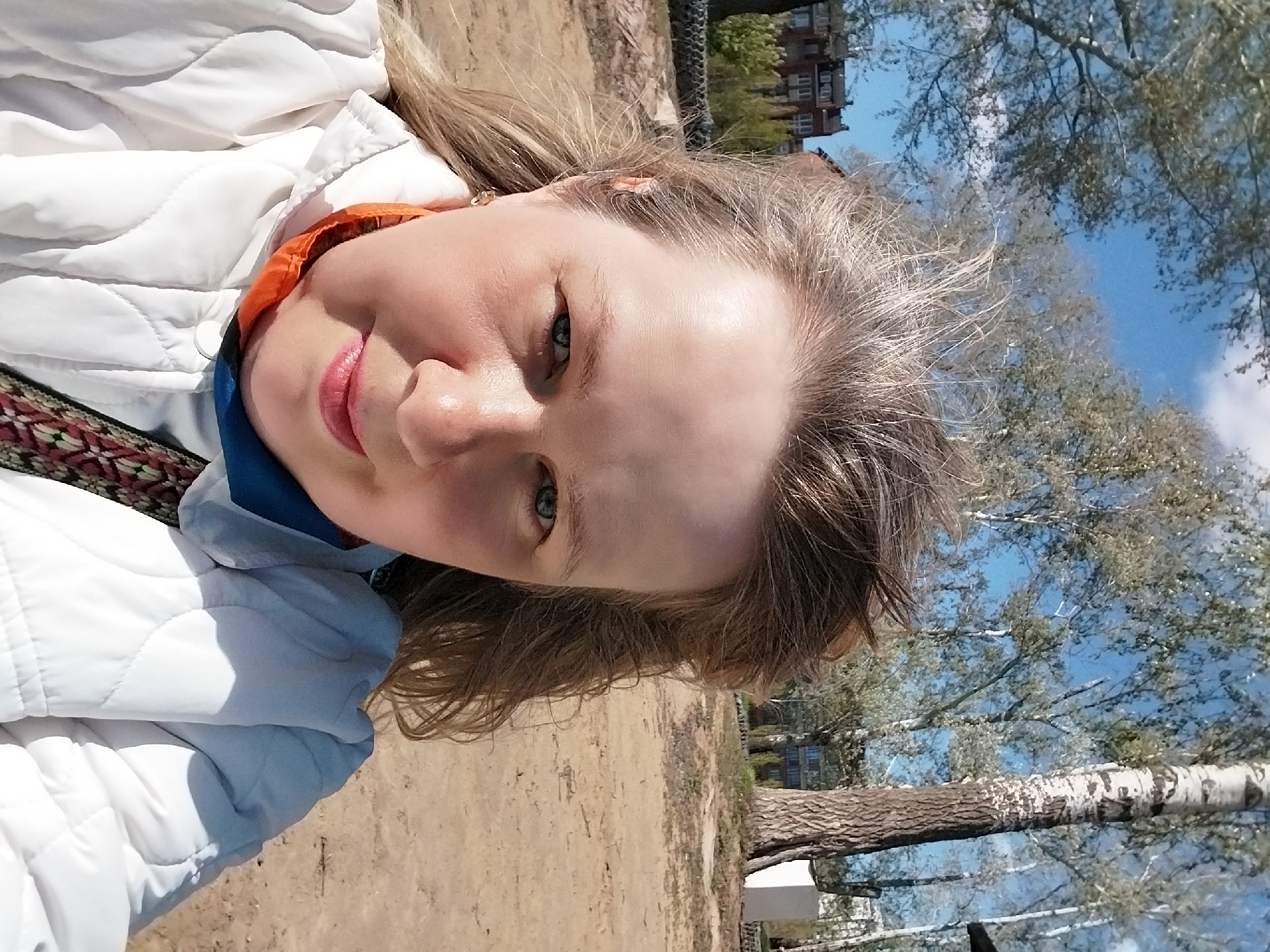 A woman is taking a selfie outside, likely in a park or open area within the city of Orenburg, Russia. She has blonde hair and fair skin, wearing a white quilted jacket with an orange and blue inner collar. The background features a dirt path, trees with budding leaves against a blue sky, and buildings in the distance. A birch tree is visible in the lower right corner.