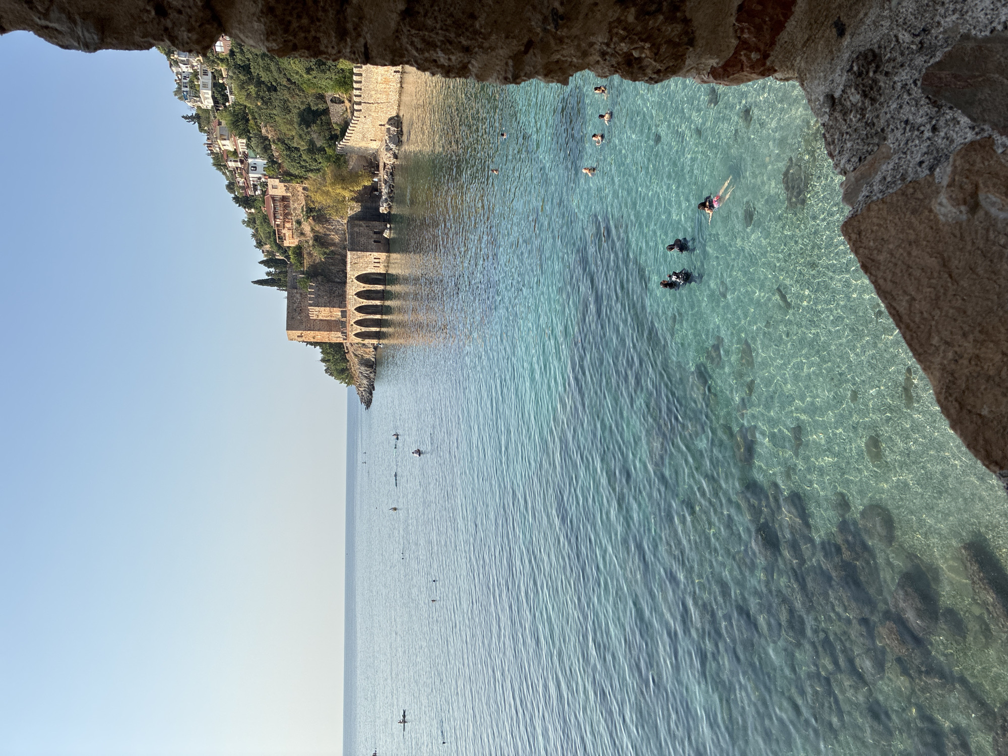 The image shows a scenic view of the Alanya Castle and the Mediterranean Sea in Alanya, Turkey. In the foreground, a stone wall from the castle's fortifications frames the right side of the image. The sea is crystal clear, revealing the rocky seabed below. Several people are swimming and snorkeling in the turquoise waters. In the middle ground, the historical Alanya Castle, with its distinctive red-brick walls and arches, stands prominently on a hilltop overlooking the sea. White houses with red-tiled roofs are scattered across the hillside, typical of the town's architecture. The sky is a clear, bright blue. The scene captures a sunny day with calm seas and recreational activities.