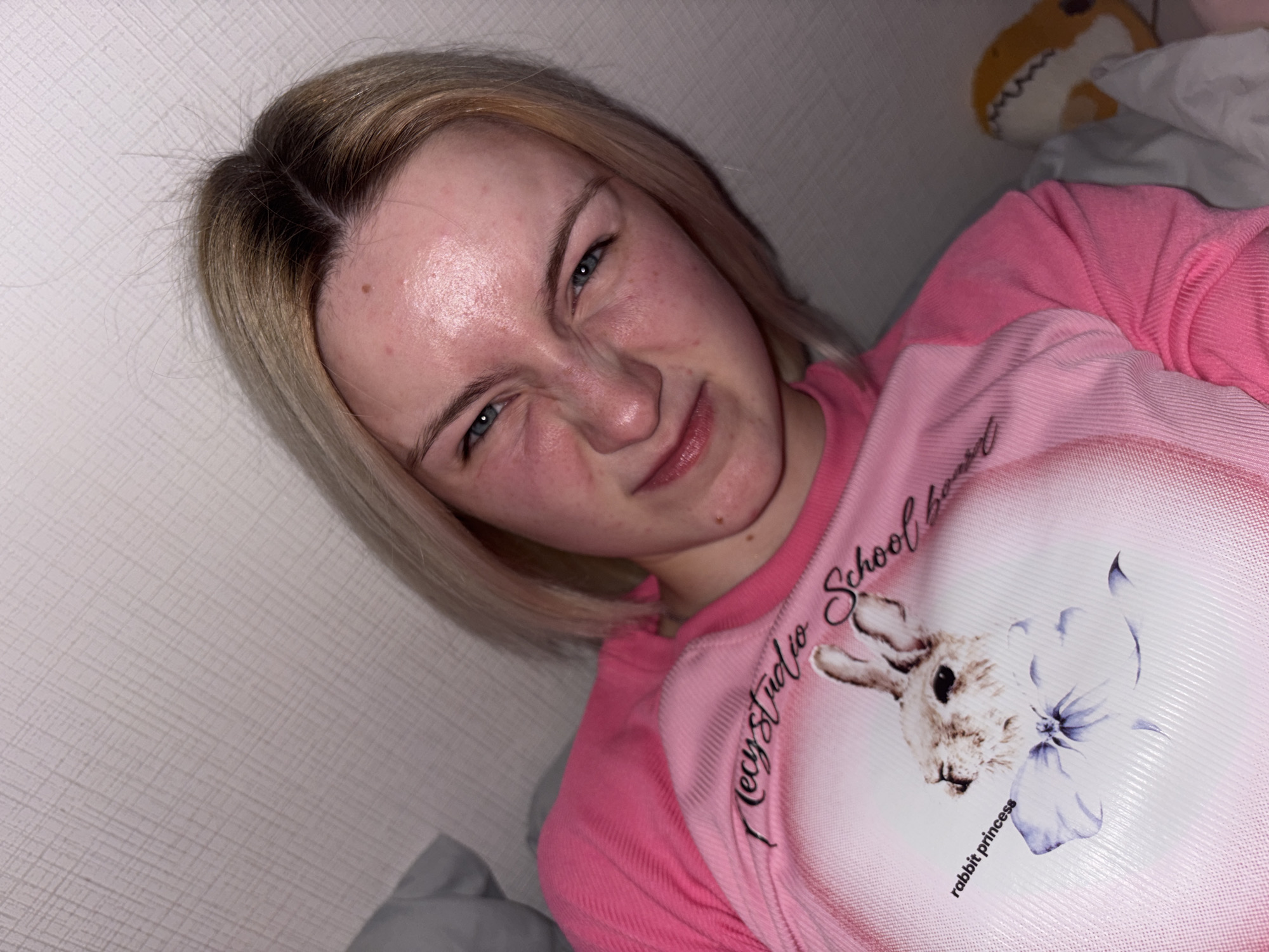 The image features a young woman, likely in her late teens or early twenties, captured indoors in Ligolambi, Russia. She has blonde hair with noticeable dark roots and blue eyes. Her skin displays some redness and minor blemishes. The woman is making a distinct scrunching or playful facial expression, narrowing her eyes and wrinkling her nose.
She is dressed in a pink long-sleeved shirt, which has a large, lighter pink oval-shaped graphic on the chest. This graphic features the head of a rabbit alongside a purple flower. Text visible on the graphic includes "Mecy Studio School" (partially obscured) and "rabbit princess."
The background suggests an indoor residential setting, possibly a bedroom, characterized by a light-colored, textured wall or headboard. In the upper right corner of the frame, a portion of a light-colored stuffed animal, which appears to be a shark with an open mouth and teeth, is visible. The lighting is artificial, illuminating her face softly. The photograph is taken from a slightly angled perspective, suggesting the woman is lying down or reclining. Her expression conveys a lighthearted and quirky mood.