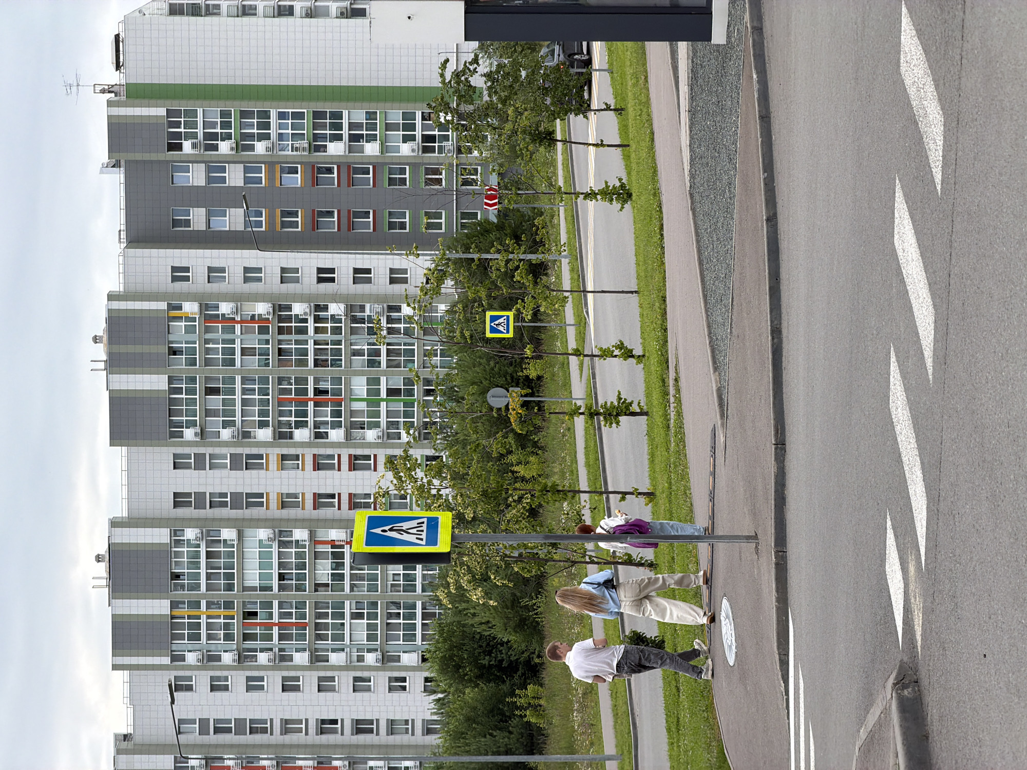 The image captures a street scene in Kazan, Russia, featuring a modern residential building. The building is tall and has a grey and white facade with green accents and numerous windows. In the foreground, a crosswalk is visible on the asphalt road, marked with white stripes. A pedestrian crossing sign, a yellow square with a blue border and a white silhouette of a person walking, stands near the crosswalk. Three individuals are depicted near the crosswalk. One person, seen from the back, is wearing a white t-shirt and dark pants. Another person, also seen from behind, is wearing a light-colored top and pants, with long blonde hair. A third person, partially visible, is wearing a purple jacket and light-colored pants. They appear to be waiting or preparing to cross the street. The presence of trees lining the sidewalk and the overcast sky suggest it might be daytime with cloudy weather. The overall setting is urban, likely a residential area given the apartment building. No discernible text is visible in the image.