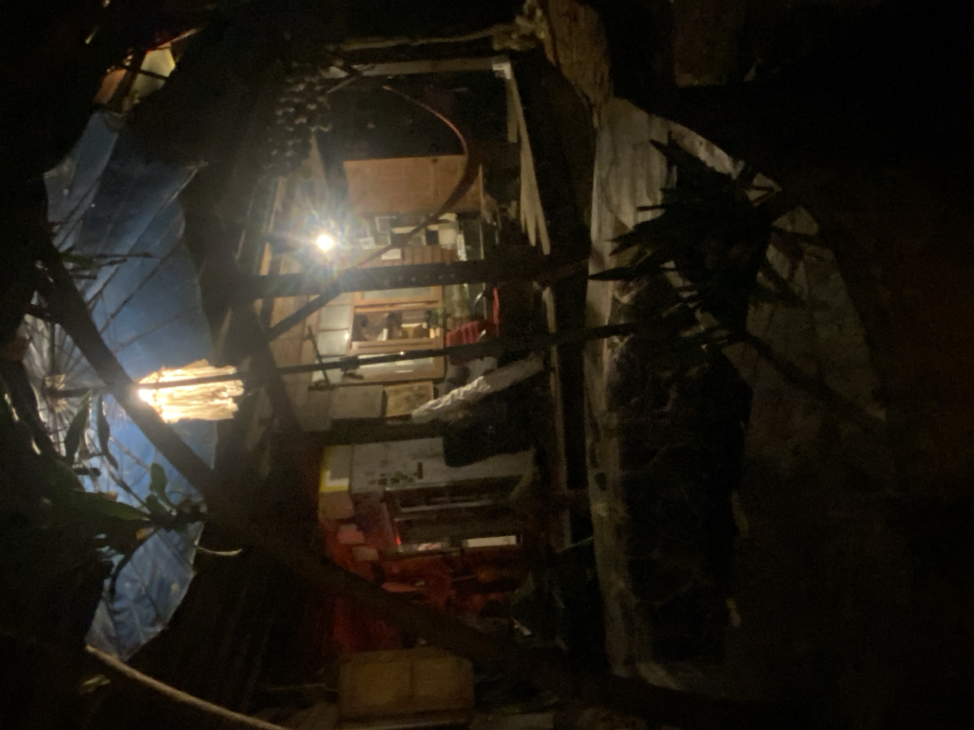 View from the exterior into the cluttered interior of a building in Baan Don Sai, Thailand. The foreground is dark, with what appear to be tree branches and foliage obscuring parts of the view. Through gaps, a cluttered interior is visible, containing furniture, boxes, and various other objects. Light emanates from within the building, illuminating some of the interior. The interior space appears to be multi-level. Overall, the scene has a chaotic and somewhat disorganized appearance.