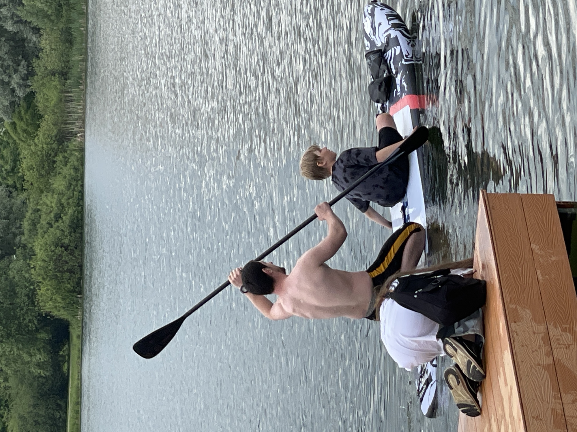 This image depicts an outdoor recreational scene on a body of water, likely a lake or wide river in Moscow, Russia, during clear daytime conditions.
In the immediate foreground, part of a light-colored wooden dock or platform is visible. On this dock, a person is partially seen from behind, wearing a white t-shirt, light-colored shorts, and dark sneakers with white soles, with a black shoulder bag. Adjacent to them, a shirtless man with dark hair and a beard, wearing black shorts with yellow stripes, is poised to engage with a stand-up paddleboard. He is holding a long, black paddle, suggesting he is either preparing to push off from the dock or actively paddling.
Seated or kneeling on the white paddleboard is a child or young person with light brown hair, wearing a dark patterned t-shirt and dark shorts. The paddleboard itself features black and white abstract designs on its front section and a prominent red stripe in the middle, with a black utility pouch attached.
The water surface is calm, showing gentle ripples and reflections. In the background, the far bank of the water body is densely covered with lush green trees and foliage, indicating a natural or park-like environment. The overall atmosphere is one of leisure and outdoor water sports activity. No visible text is present in the image.