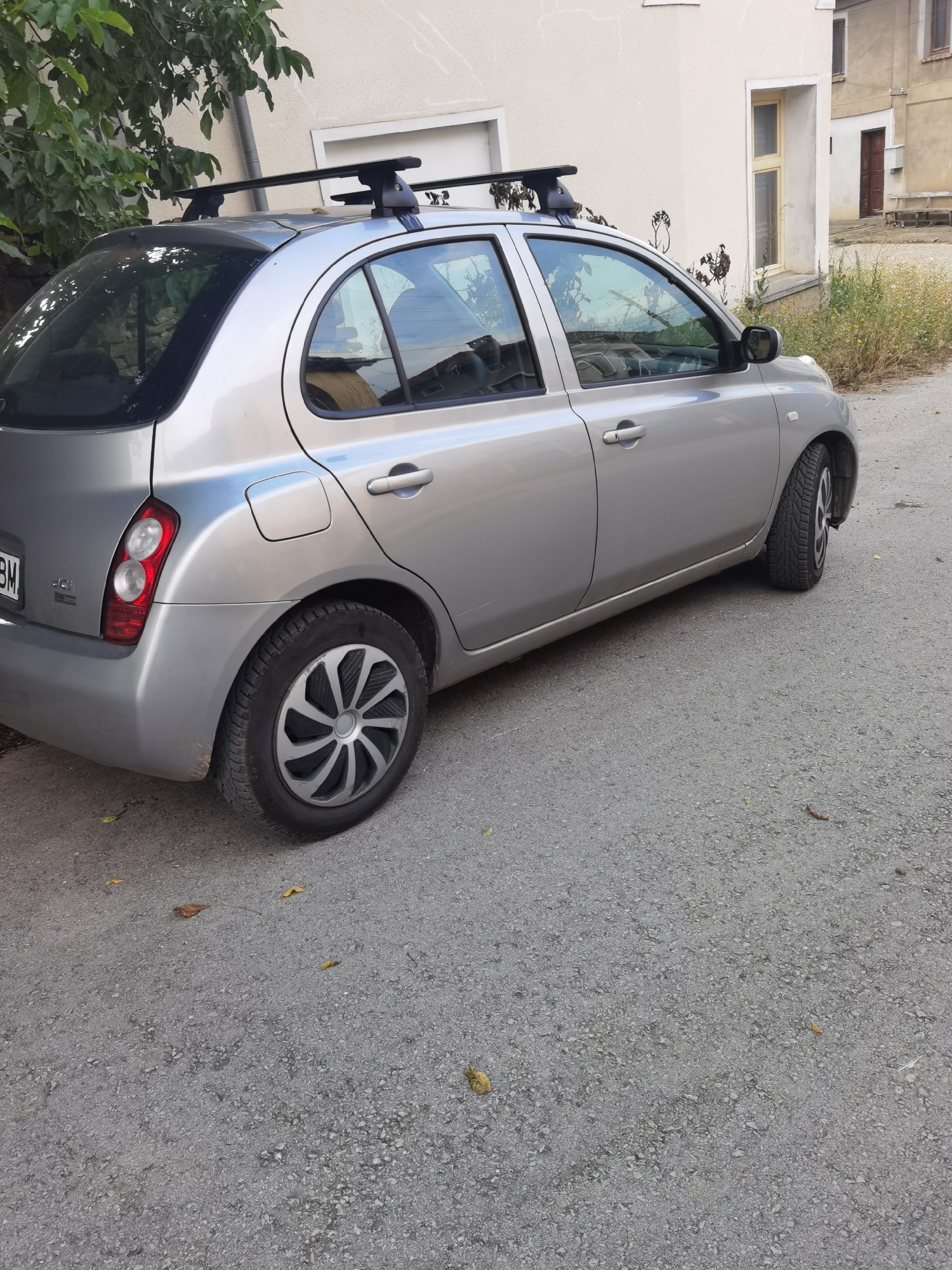 A silver Nissan car is parked on a paved street in front of a building. The car has a roof rack. The street is asphalt and there are some fallen leaves on the ground. The building appears to be old, with light-colored walls and windows. The background shows more buildings and some vegetation. The image was taken in Lomets, Bulgaria.