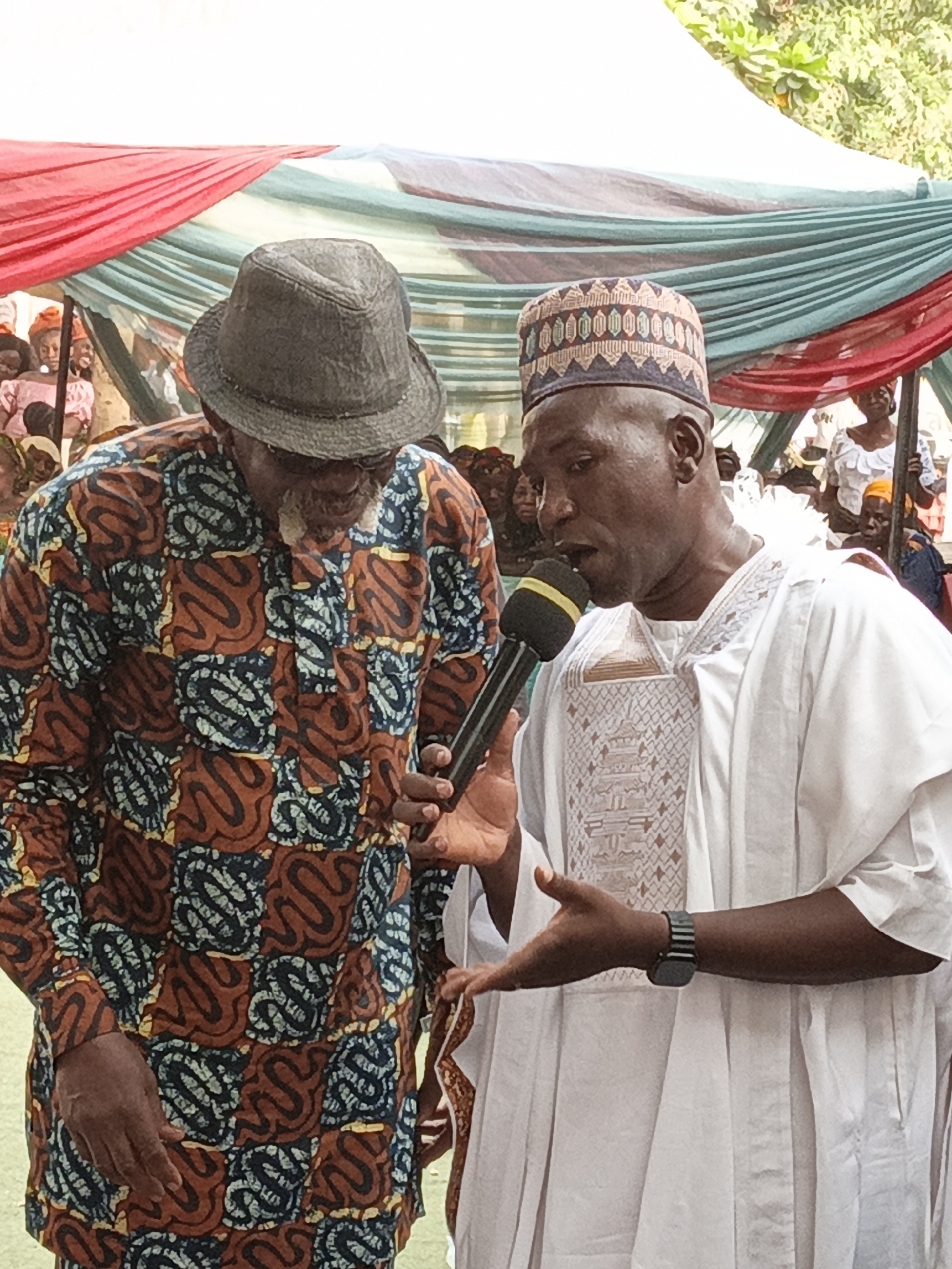 The image captures two men in traditional attire at an outdoor event in Sabon Gari West, Nigeria. In the foreground, a man on the right, with dark skin, wears a white embroidered traditional garment and a patterned cap. He holds a black microphone with a yellow band close to his mouth, appearing to speak or perform, with his right hand gesturing. A dark watch is visible on his left wrist.
To his left, an older man with a beard, wearing a dark fedora-style hat, leans in towards the microphone, looking down. He is dressed in a vibrant, long-sleeved patterned shirt featuring warm earthy tones of brown, orange, and yellow, interspersed with blue and black designs. His dark-skinned hand is visible on his left side.
The setting is an outdoor gathering, indicated by the draped fabric canopy overhead, which features red, light blue, and possibly other colors. The background is filled with numerous blurred figures of people, suggesting a crowd or audience. Several children's faces can be seen peeking from behind the canopy fabric. The bright natural lighting indicates it is daytime. The traditional clothing worn by the men and the general gathering atmosphere point to a community event, possibly a celebration or public address. There is no legible text visible in the image.
