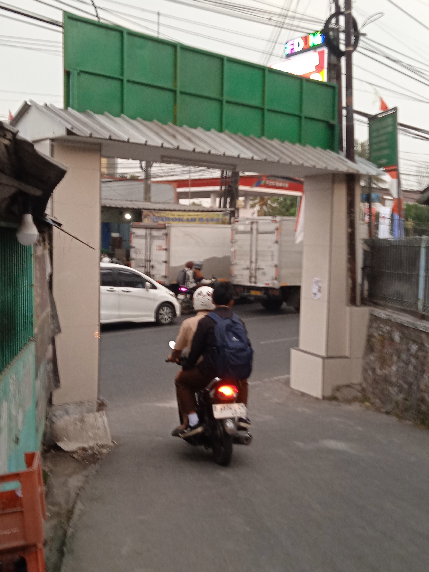 In Cileunyi, Indonesia, on a cloudy day, two individuals are riding a motorcycle past a large, arched gateway. The motorcycle, a dark color with a license plate, has two occupants. The person in the back is wearing a dark backpack, and the person in front has a white helmet. They are moving away from the viewer, towards a road where a white van and a large white truck are visible in the background. The truck appears to be stationary. The gateway is made of light-colored pillars supporting a corrugated metal roof. To the left of the gateway, a green metal fence is visible, and a single light bulb hangs from an overhang. Above the scene, a large green billboard is mounted on a structure, and a neon sign with colorful lights is attached to a pole. Flags are also visible in the background, one on a pole to the right of the neon sign and another attached to the truck. The road surface is asphalt, and there is a stone wall on the right side of the image.