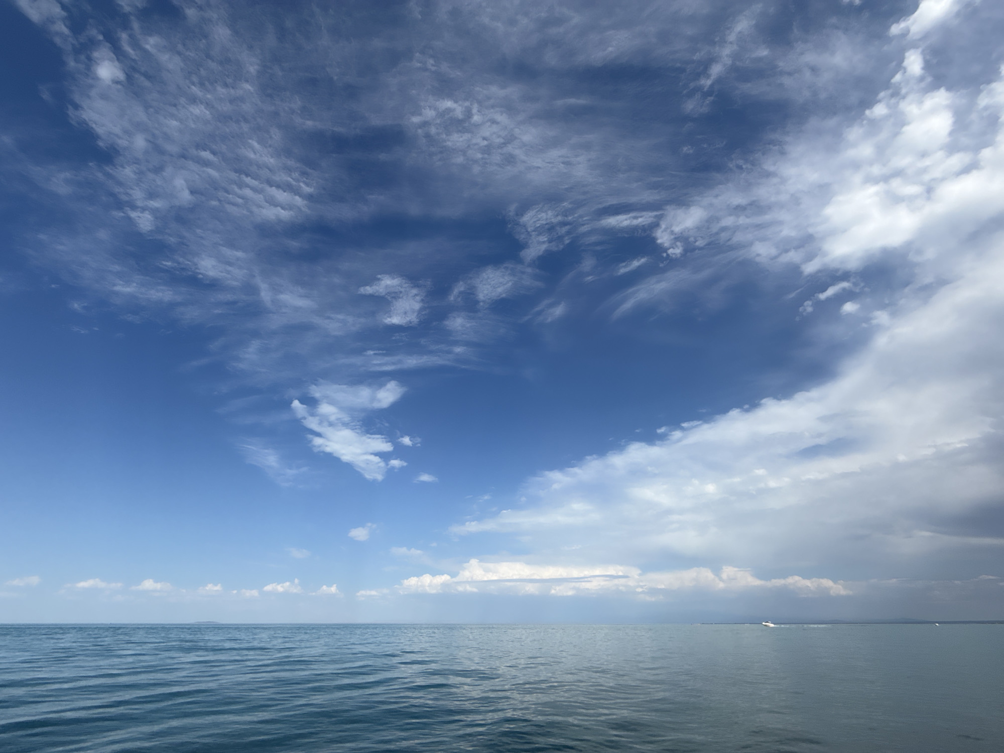 The image depicts a wide expanse of the Kakanar Lake near the village of Ақши, Kazakhstan. The sky is a vibrant blue, streaked with wispy white clouds that create a dynamic texture. The water of the lake is calm, with gentle ripples reflecting the sky. In the distance, a faint outline of land can be seen on the horizon, and a small white boat is visible moving across the water. The overall scene is one of natural beauty and tranquility.