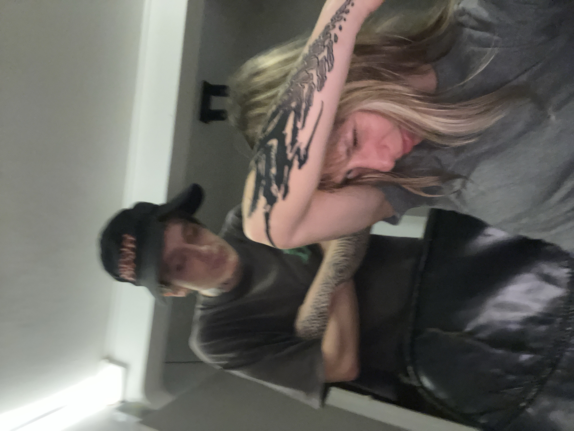 This image captures two individuals in an indoor setting. On the right, a person with long, light brown hair is shown resting, possibly asleep, with their head tilted downwards. They are wearing a grey top, and a prominent dark tattoo, featuring abstract or skeletal designs, covers a large portion of their right arm.
To their left, another individual is wearing a dark t-shirt and a dark baseball cap. The text "NSF" or similar letters are visible in a lighter color on the front of the cap. This person also has extensive tattoos on their left arm, appearing to be full sleeves with intricate patterns that resemble scales or reptilian textures. Their face is partially obscured by the cap and the angle, making their expression unclear. They appear to be standing or sitting close to the first person, with their arms crossed.
The background consists of a plain light-colored wall or ceiling, with a dark, indistinct object possibly mounted on the wall. A dark, possibly quilted material (like a blanket or jacket) is visible in the lower foreground. The lighting is subdued, suggesting an indoor environment, potentially in the evening or at night. The overall scene conveys a sense of quiet intimacy or relaxation. The context provided indicates the location is Vicente López, Argentina.