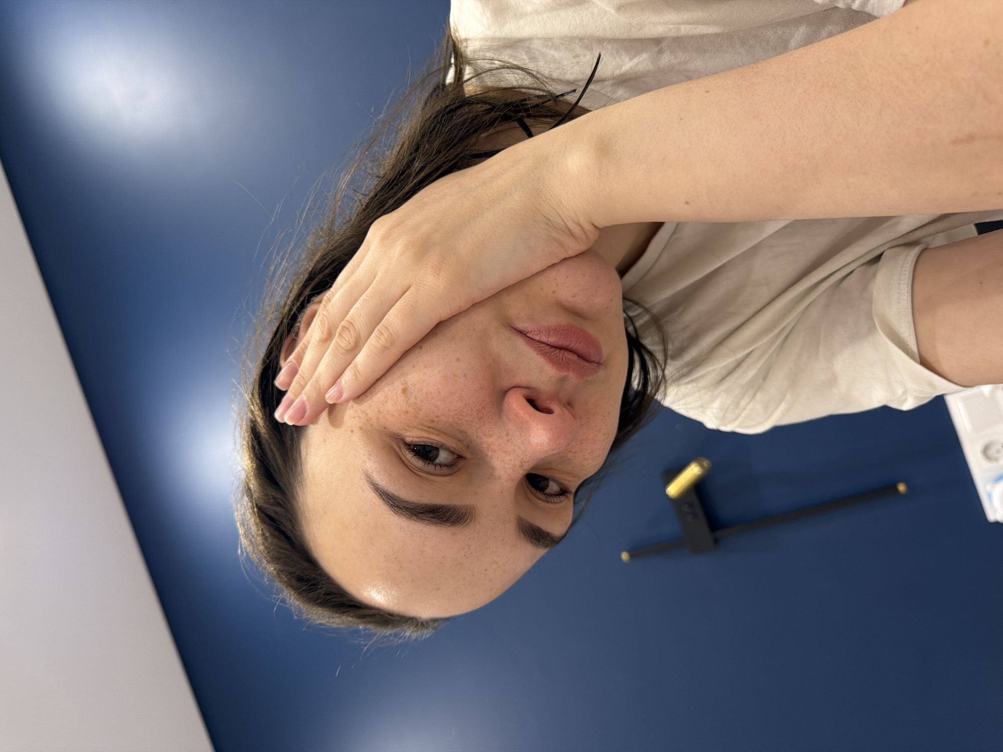 A woman with dark hair and freckles is facing the camera, with her right hand partially covering her face. She is wearing a light-colored t-shirt. The background is a dark blue wall with a black and gold decorative element resembling a clock hand. In the upper right corner, there's a glimpse of a white object. The picture does not provide enough context to verify location within Chaikovsky, Russia.