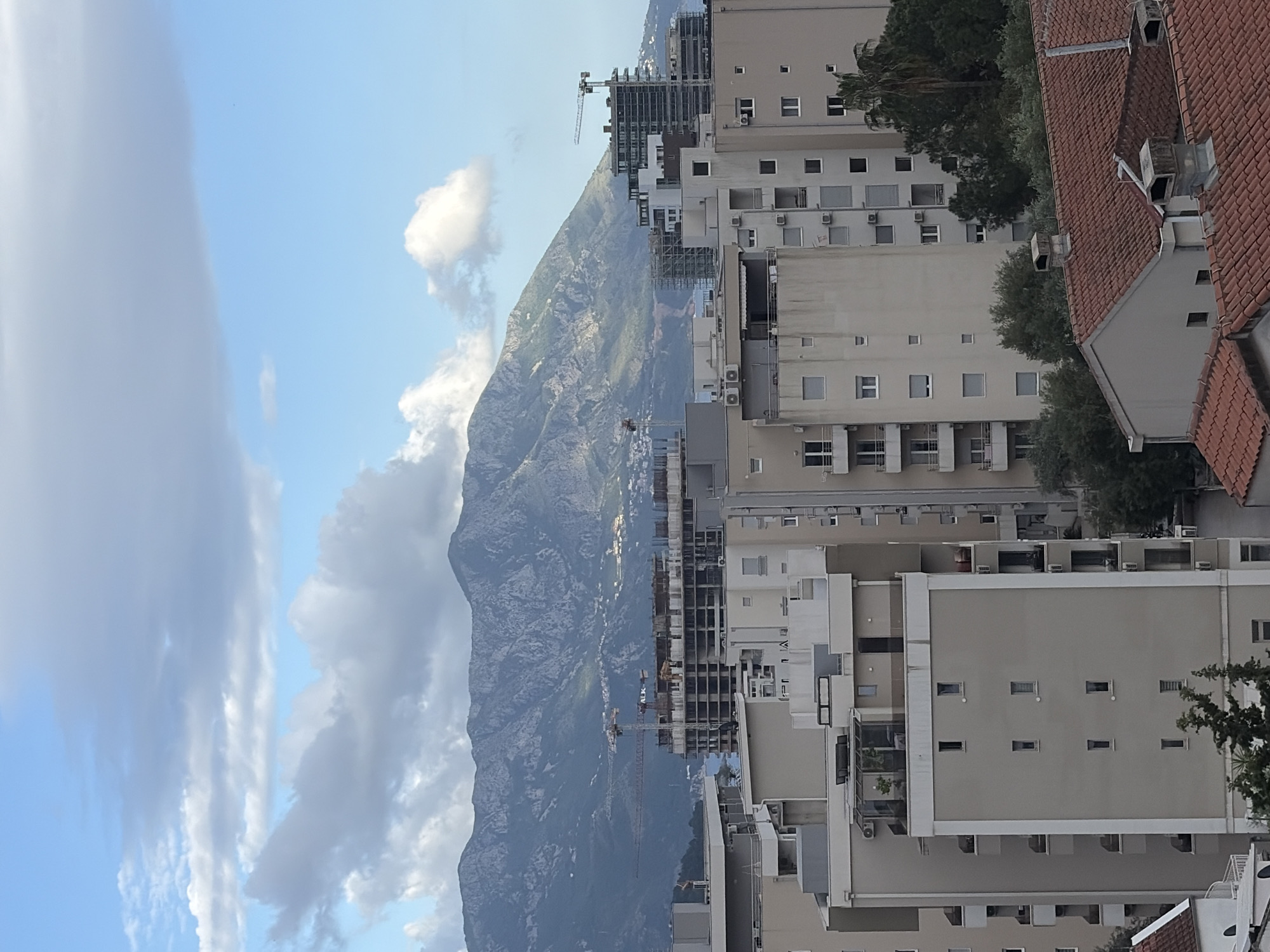 The image captures a view in Budva, Montenegro, with a mountain range forming the backdrop. In the foreground, numerous tall, beige-colored apartment buildings dominate the urban landscape, some of which are under construction, evidenced by scaffolding and cranes. The sky is partly cloudy, with shades of blue visible. A section of a building with a red tile roof is seen on the right side of the frame. Trees are interspersed among the buildings.