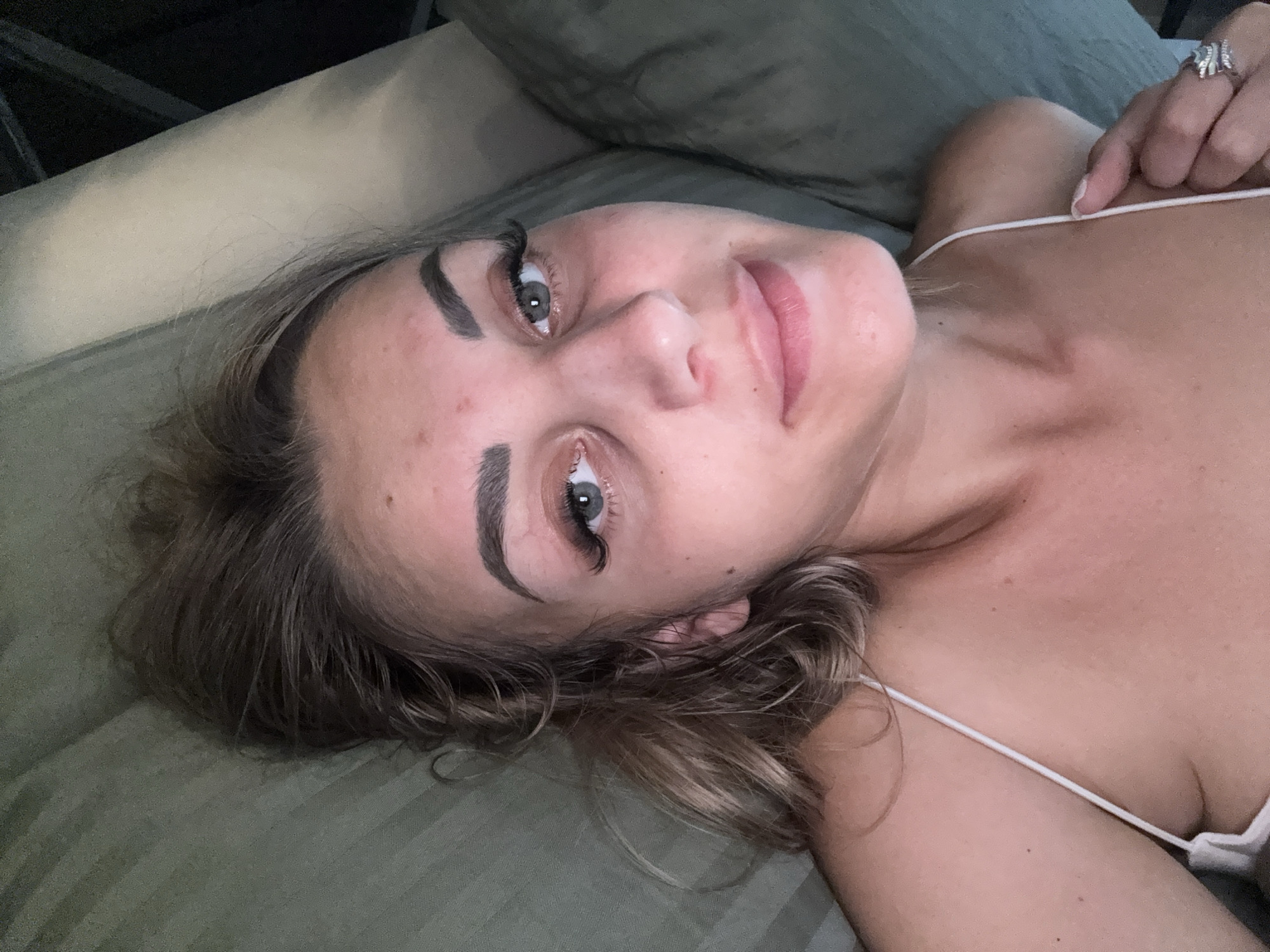 The image is an extreme close-up of a woman lying down. She has light brown hair, dark eyebrows, and light-colored eyes. Her face is slightly angled towards the camera. She is wearing a white spaghetti strap top and a ring on her right ring finger. The background appears to be a textured surface, possibly a couch or bed, in a muted green color. The lighting is dim, suggesting an indoor setting. The location in Yuzhno-Sakhalinsk, Russia, is not discernible from the image.