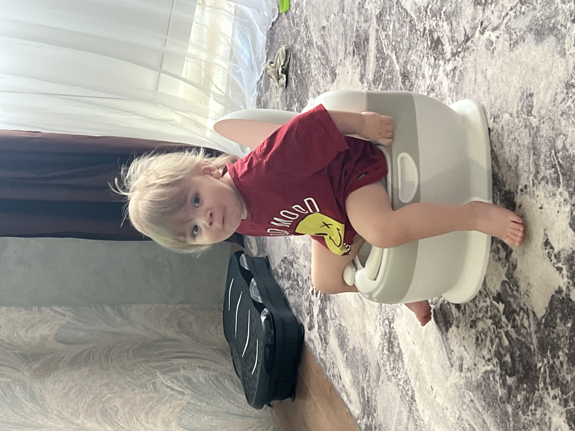 A young child with blond hair, wearing a red t-shirt with a yellow smiley face, is sitting on a white and grey potty chair on a rug with a grey and white marbled pattern. The child is looking towards the viewer. The background includes a wall with a patterned design, a dark brown curtain, a white sheer curtain, and a window. A black scooter is leaning against the wall. A discarded clothing item can be seen in the background. The photo was taken indoors, possibly in a residential setting. There is no verifiable information indicating the location of the image is in the city of Noviy, Russia.
