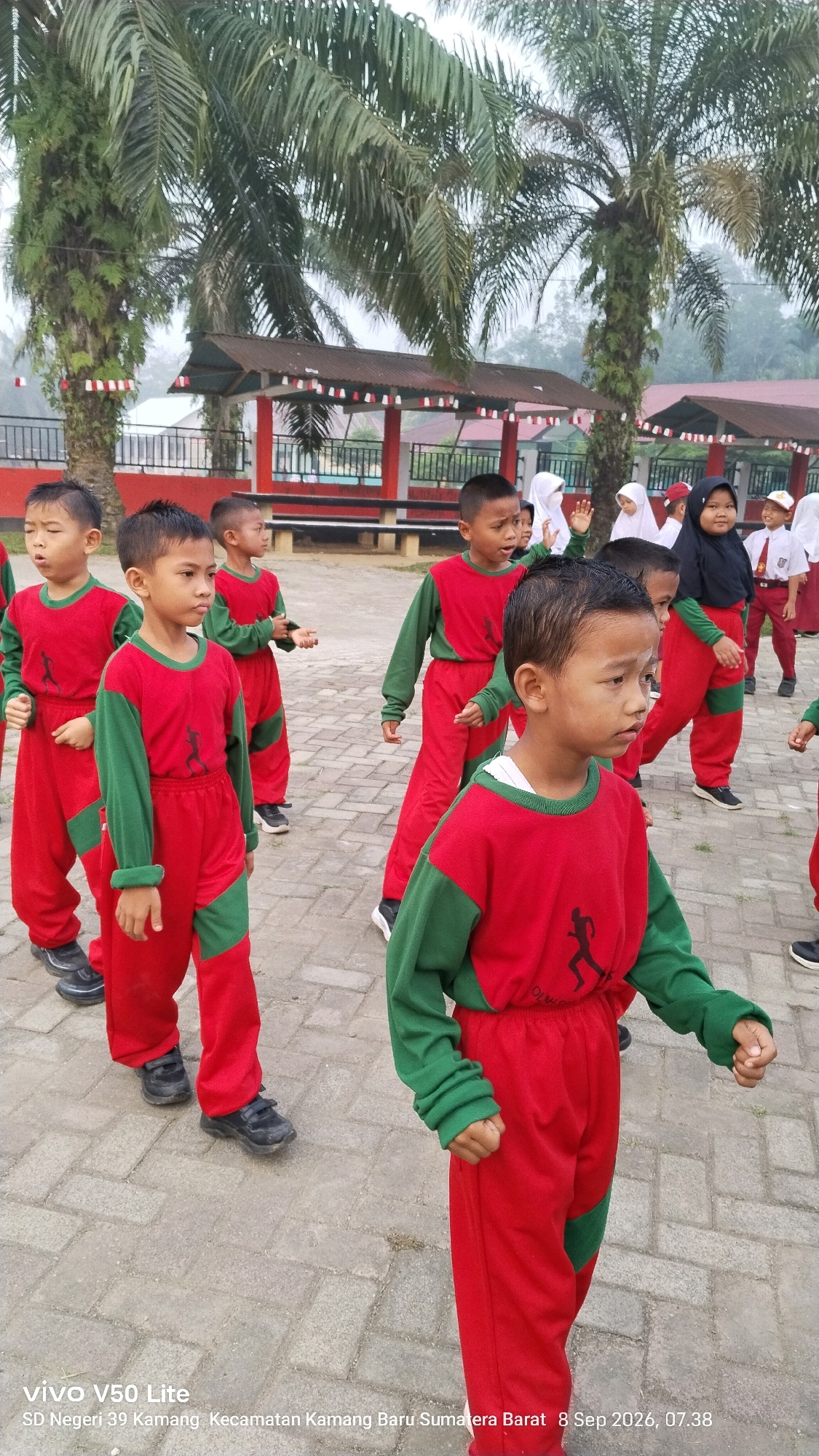In an outdoor scene, several young children, appearing to be of elementary school age, are gathered on a paved area. They are all dressed in matching red and green athletic wear, with the boys wearing long-sleeved tops and pants, and some of the girls in the background wearing hijabs and school uniforms. The children are facing in various directions, with some looking towards the camera and others engaged in some activity. In the background, there are palm trees, a building with red and white trim, and a covered seating area. The overall atmosphere suggests a school event or activity, possibly a physical education class or a school gathering. The bottom of the image includes text overlay indicating "vivo V50 Lite" and "SD Negeri 39 Kamang, Kecamatan Kamang Baru, Sumatera Barat, 8 Sep 2026, 07.38". This information places the scene in Sumatra Barat, Indonesia.