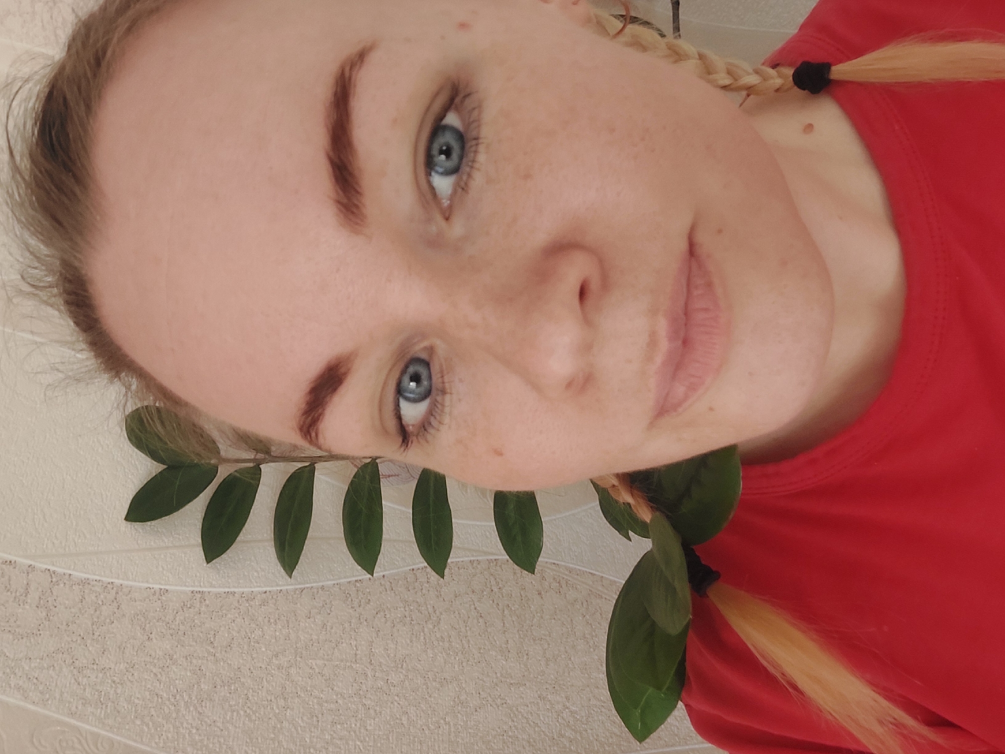 A photograph, rotated clockwise approximately 90 degrees, shows a woman's face and part of her upper body in an indoor setting. She has fair skin, freckles, and light blue eyes. Her eyebrows are reddish-brown. Her blonde hair is styled into two braids tied with black bands. She is wearing a red t-shirt. A branch with green leaves is positioned near her face, appearing to be part of the composition. The background consists of a textured, off-white wall. There is no discernible indication of the location of Большой Букор, Russia.
