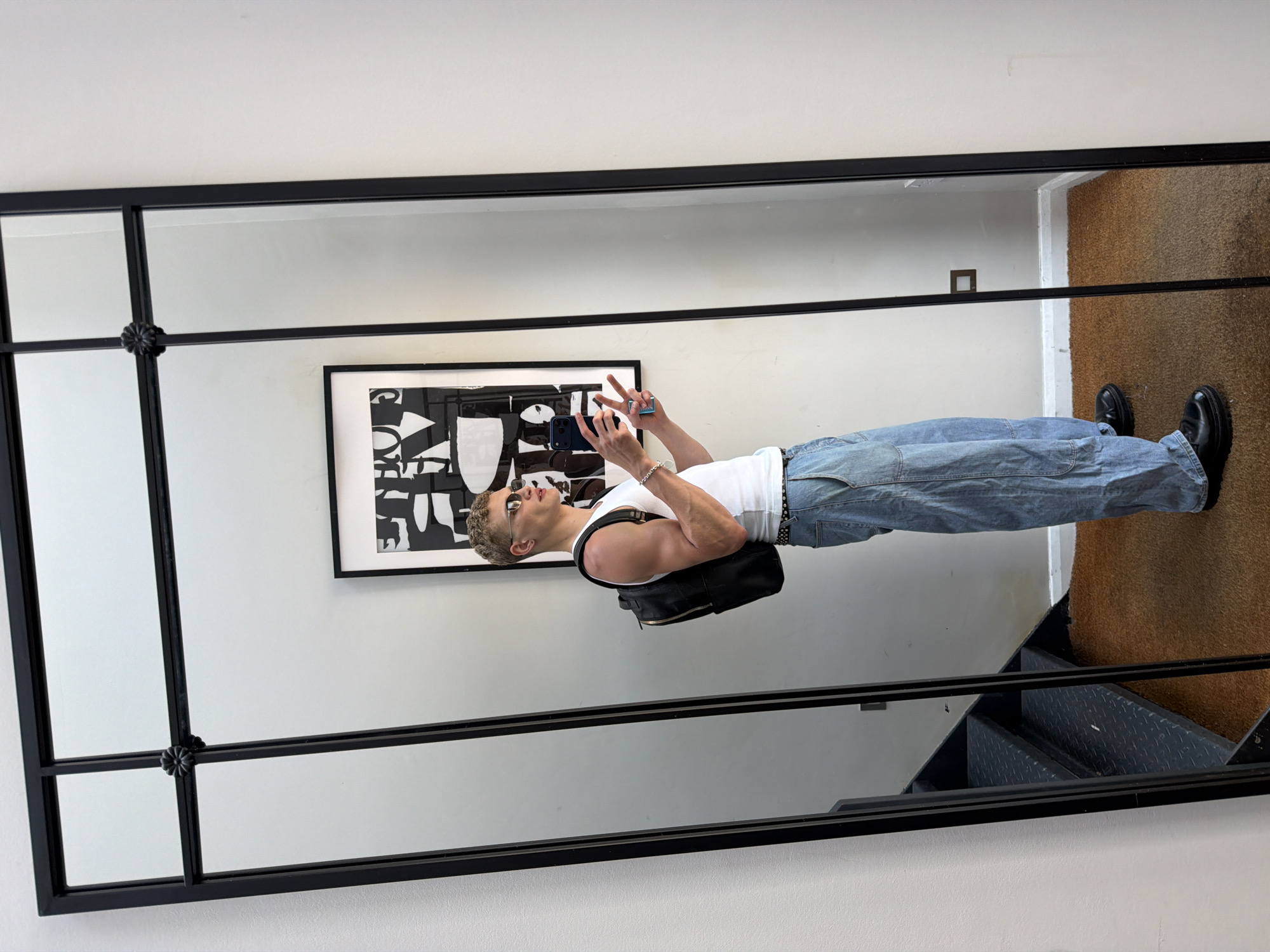 The image shows a man's reflection in a large black-framed mirror. He's fair-skinned, wearing sunglasses, a white tank top, loose blue jeans with a black studded belt, and black shoes. He is holding a phone, making a peace sign with his fingers. He also has a black backpack on. The mirror reflects a room with white walls. Behind the man's reflection is a black and white abstract artwork hanging on the wall. The floor is covered with brown matting. The bottom right corner of the mirror reflects a staircase with metal steps. The location is likely in London, United Kingdom, based on architectural styles and interior design.