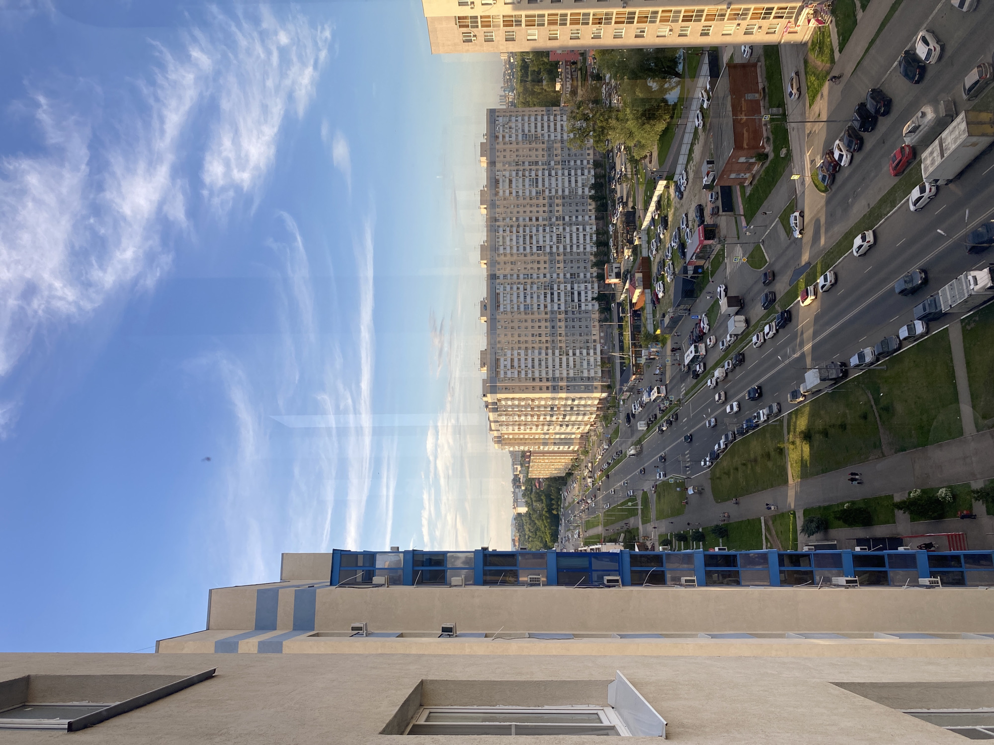 The image depicts an urban scene in Novokuybyshevsk, Russia, captured from an elevated viewpoint, likely a high-rise building. In the foreground on the left, the beige exterior of a modern apartment building is visible, with a white-framed window and a white windowsill. Adjacent to it is another building with a facade featuring alternating bands of beige and light blue, with multiple floors of blue-tinted balconies and windows.
The main focus of the image is a wide, multi-lane road running horizontally across the mid-ground, lined with trees and sidewalks. Traffic is moderately heavy, with numerous cars, trucks, and a bus visible. The vehicles are moving in both directions. On the right side of the road, a long, light-beige apartment building with numerous windows stretches across the frame. Further down the road, a much larger, ten-story apartment building with a patterned facade is prominent. The background reveals more buildings and a dense canopy of green trees, suggesting a residential area.
The sky above is a clear blue with wispy white clouds, indicating fair weather. The sunlight illuminates the buildings, casting long shadows and creating a bright, airy atmosphere. Several people are visible walking on the sidewalks and near the buildings, and some are seen near parked cars. The overall impression is of a typical day in a modern Russian city.
