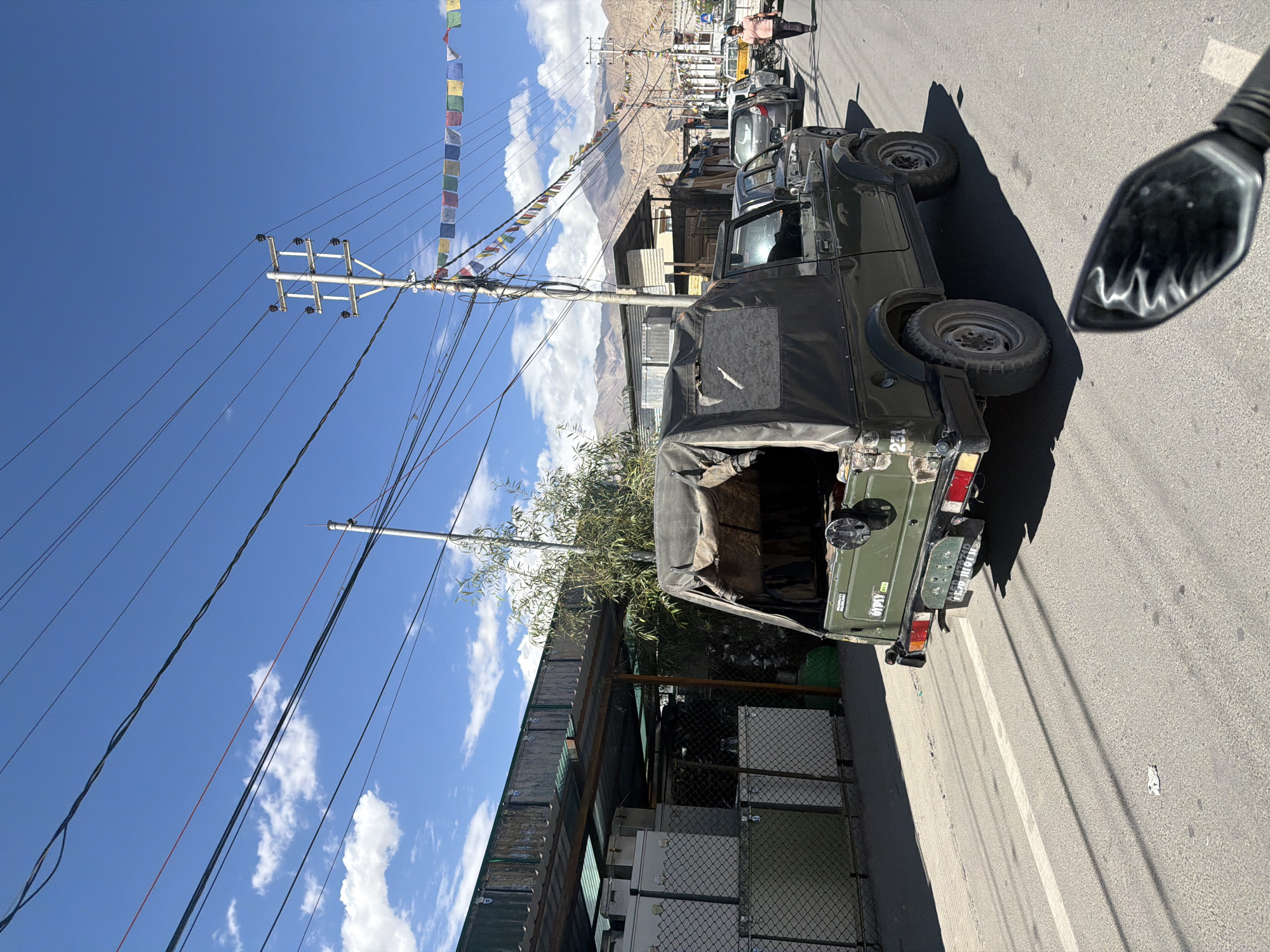 In the city of Leh, India, a dark green Suzuki Gypsy vehicle, with a canvas top and military-style markings, is parked on the right side of a paved road. The vehicle displays license plates and a number "231" on its side. The road is bordered by a sidewalk on the left, where white, boxy structures with wire mesh fences are visible, along with a covered awning. Further down the road, several other vehicles are parked or in motion, including a silver SUV, a white hatchback, and a yellow pickup truck. A man in a pink shirt walks on the right side of the road, while a woman in a dark shirt and pants cycles further ahead. The scene is set against a backdrop of buildings and mountains under a bright blue sky with scattered white clouds. Overhead, numerous electrical wires and a utility pole with prayer flags strung across it are prominent features.