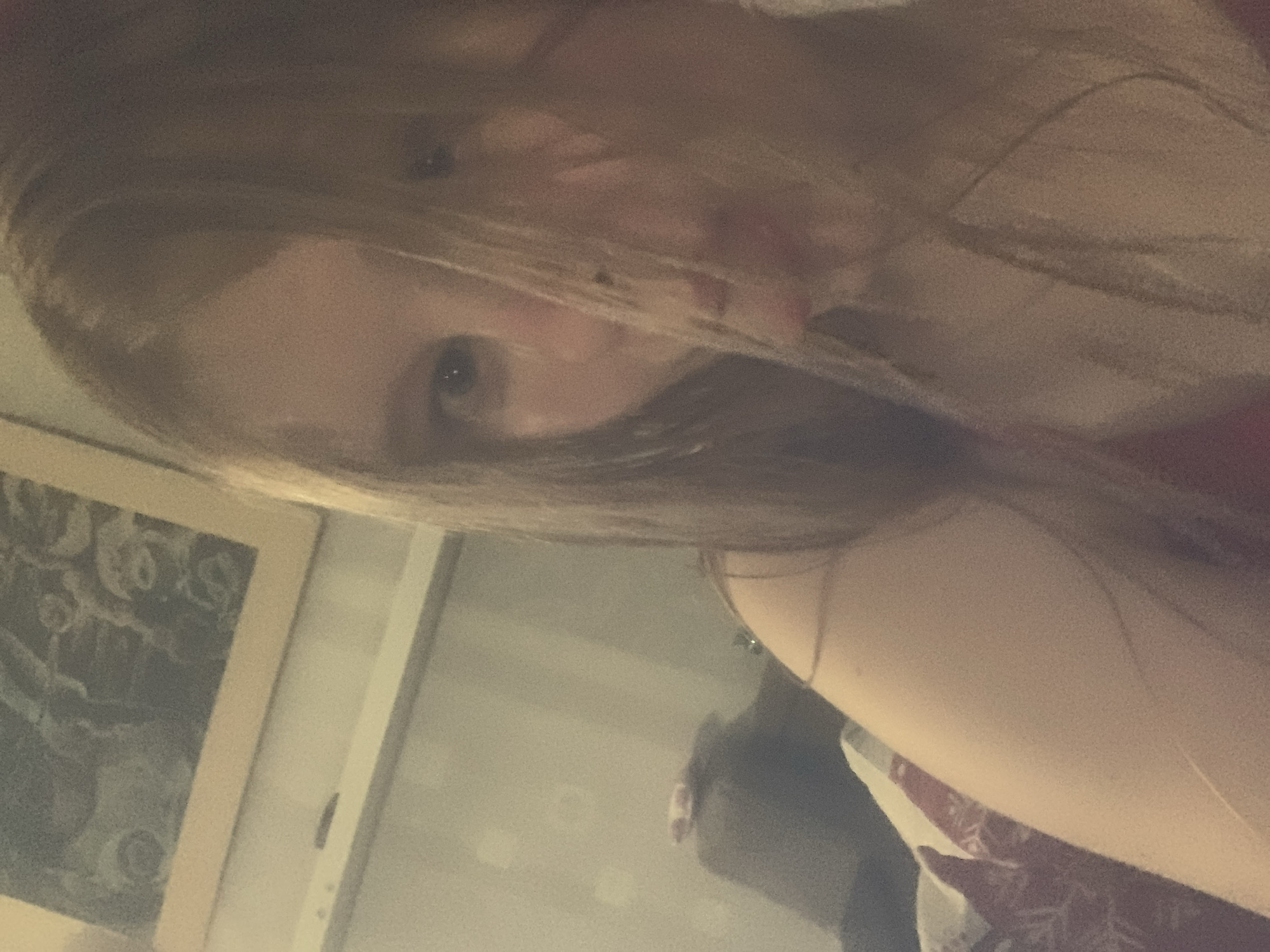 The image is a close-up, slightly blurry, indoor selfie of a young person with long, light brown hair that partially obscures their face. They have blue eyes and are looking towards the camera. The person is wearing a red garment. The background appears to be a room with a framed picture on the wall and some furniture. The overall lighting is dim. The image does not provide any information about the location in the city of Unknown, Russia.