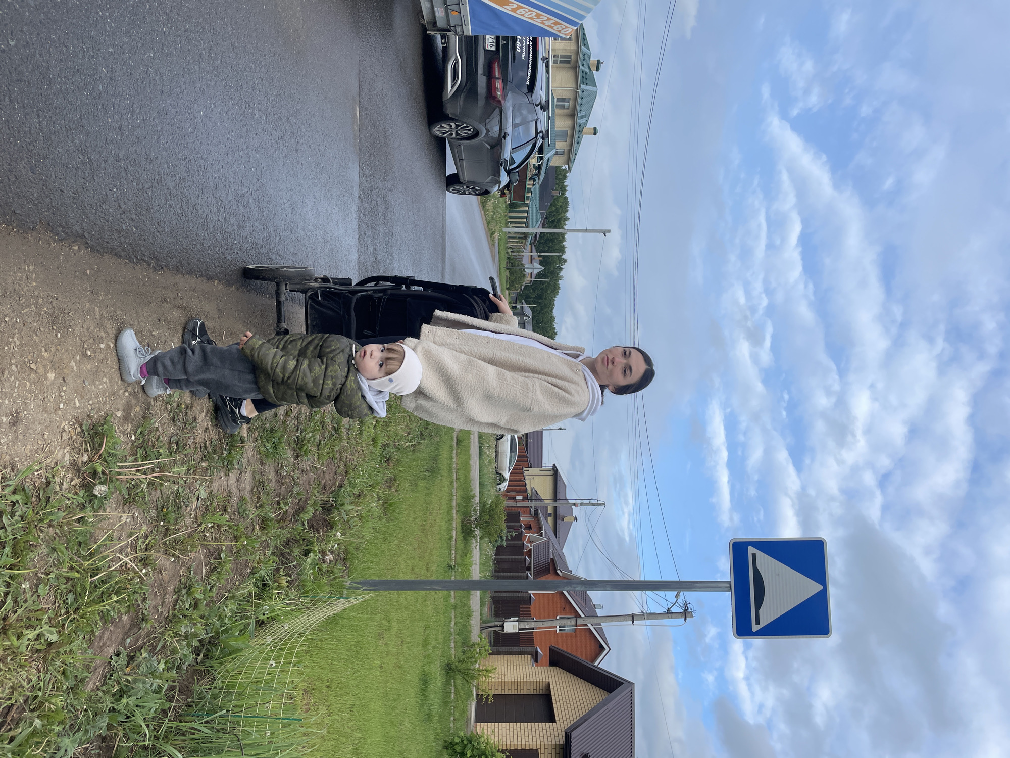 The image shows a woman with a child in a stroller standing next to a road in Эстачи, Russia. The road is wet, suggesting it has recently rained. A row of houses is visible on the other side of the road. A road sign indicating a speed bump is visible near the woman and child. Multiple cars are parked on the roadside, with a trailer attached to one of them. The sky is cloudy.