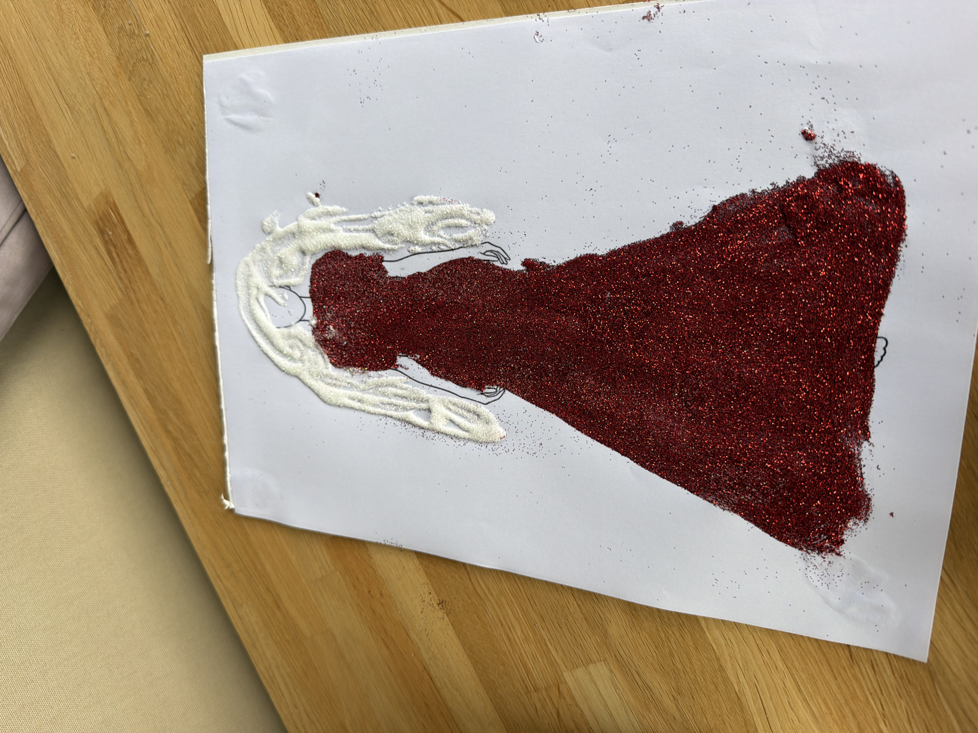 An artistic representation of a woman in a red glitter dress and white glitter hair is displayed on a white sheet of paper, which is situated on a wooden table. The drawing is outlined in black. The image appears to be a close-up shot, with the top-left portion of the frame showing a diagonally-oriented surface, possibly a wall or ceiling, with a textured pattern. The wooden table has visible grain and appears to be made of light-colored wood planks. Scattered red and white glitter particles are present on the table and the white paper, indicating the crafting process. The image does not provide any information that can be used to determine its location in Baloži, Latvia.