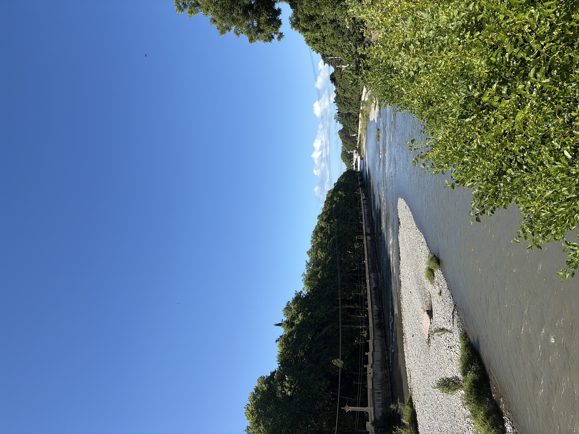 The image captures a serene view of the Mzymta River in Sochi, Russia, on a clear day. The river, with its gently flowing, muddy water, curves through a landscape flanked by lush green trees on both banks. A pebble-strewn shore is visible on the left, with a few small trees and patches of grass dotting the area. On the right bank, dense foliage creates a vibrant green barrier, partially obscuring the view of buildings and further vegetation beyond. A stone embankment with decorative pillars lines the left side of the river, suggesting a developed area, possibly a promenade or park. In the distance, a white bridge spans the river, leading towards more trees and buildings under a bright blue sky dotted with a few wispy white clouds. The overall scene conveys a sense of tranquility and natural beauty within an urban setting.