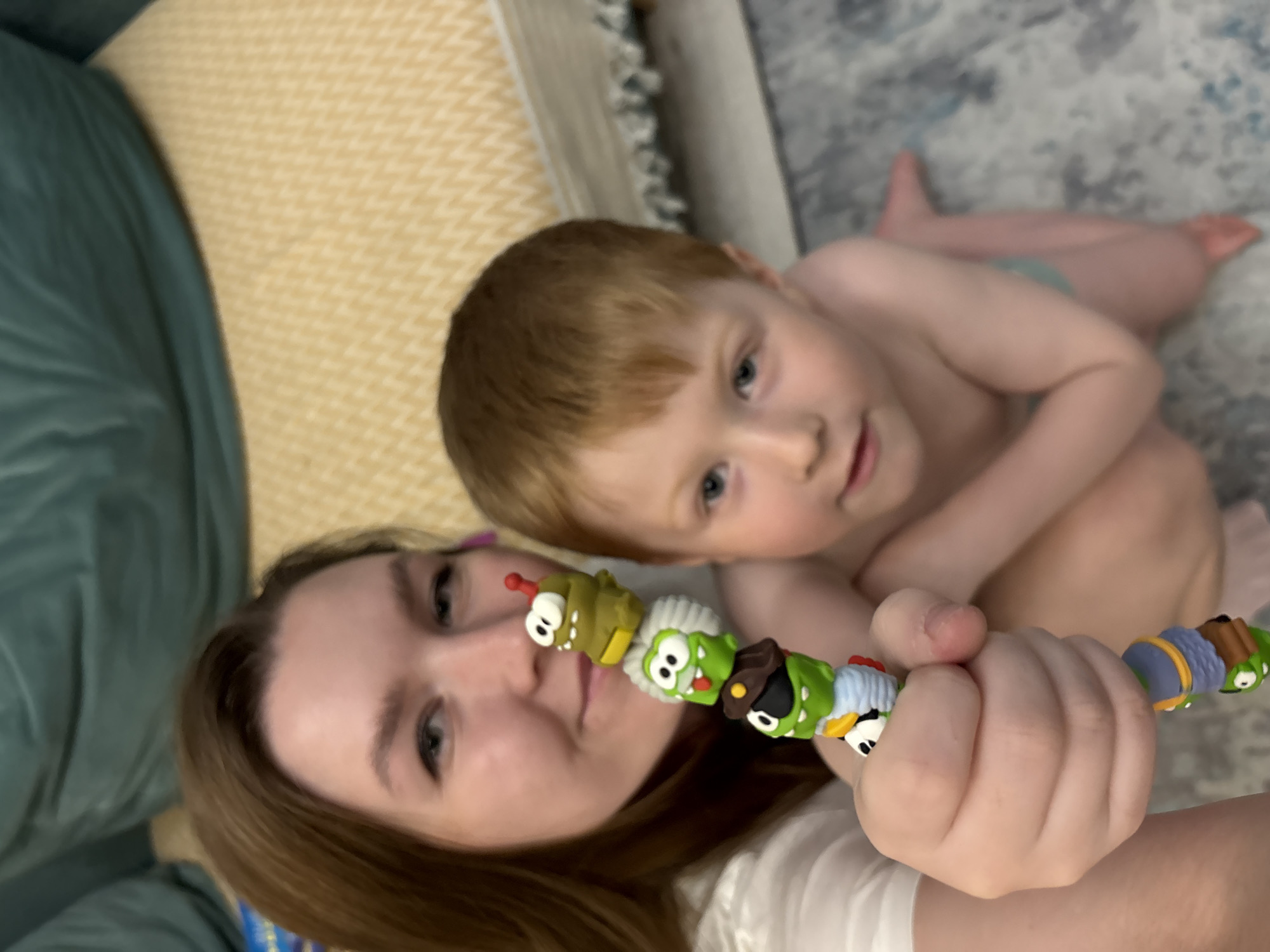 A selfie shows a woman and a shirtless young boy on what appears to be a patterned gray, blue, and white bedspread. The woman holds a colorful caterpillar toy in front of her face. The boy leans in close to the woman. The background includes a patterned beige and white pillow, and a dark green cushion or pillow. The location is not specified, and there is no information to suggest the city of Izhevsk, Russia.