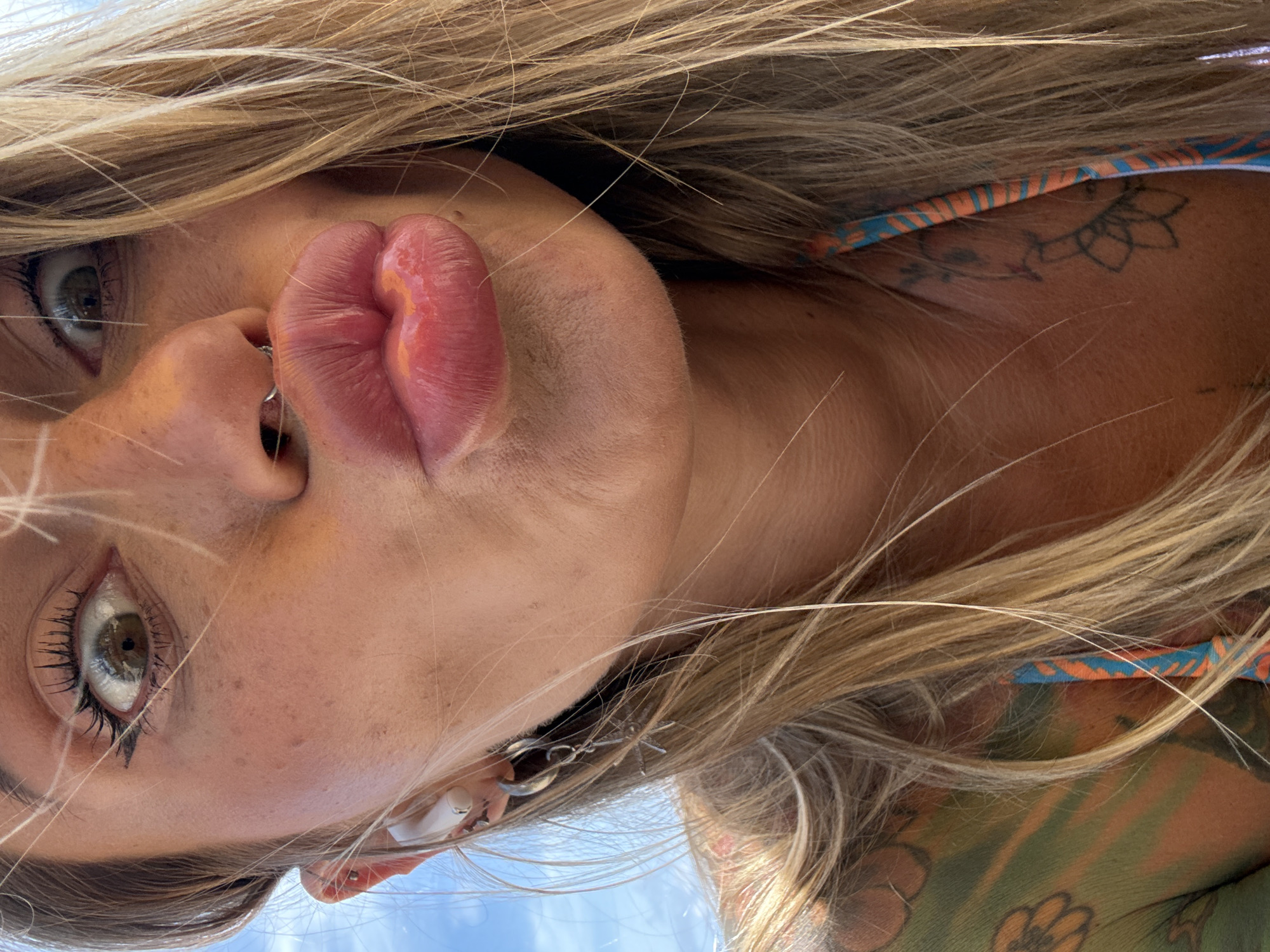 A close-up, low-angle, outdoor, full-frame shot shows the face of a blonde woman with a nose ring and ear piercings. She is puckering her lips towards the camera. Her eyes are wide open, looking directly at the camera. She is wearing a bikini top with blue and orange stripes. Her right shoulder and chest are visible, showing a colorful tattoo of flowers and other designs in green, orange, and yellow. Her blonde hair is blowing in the wind. The background consists of a bright blue sky. The scene appears to be the beach or poolside in Choeng Thale, Thailand.