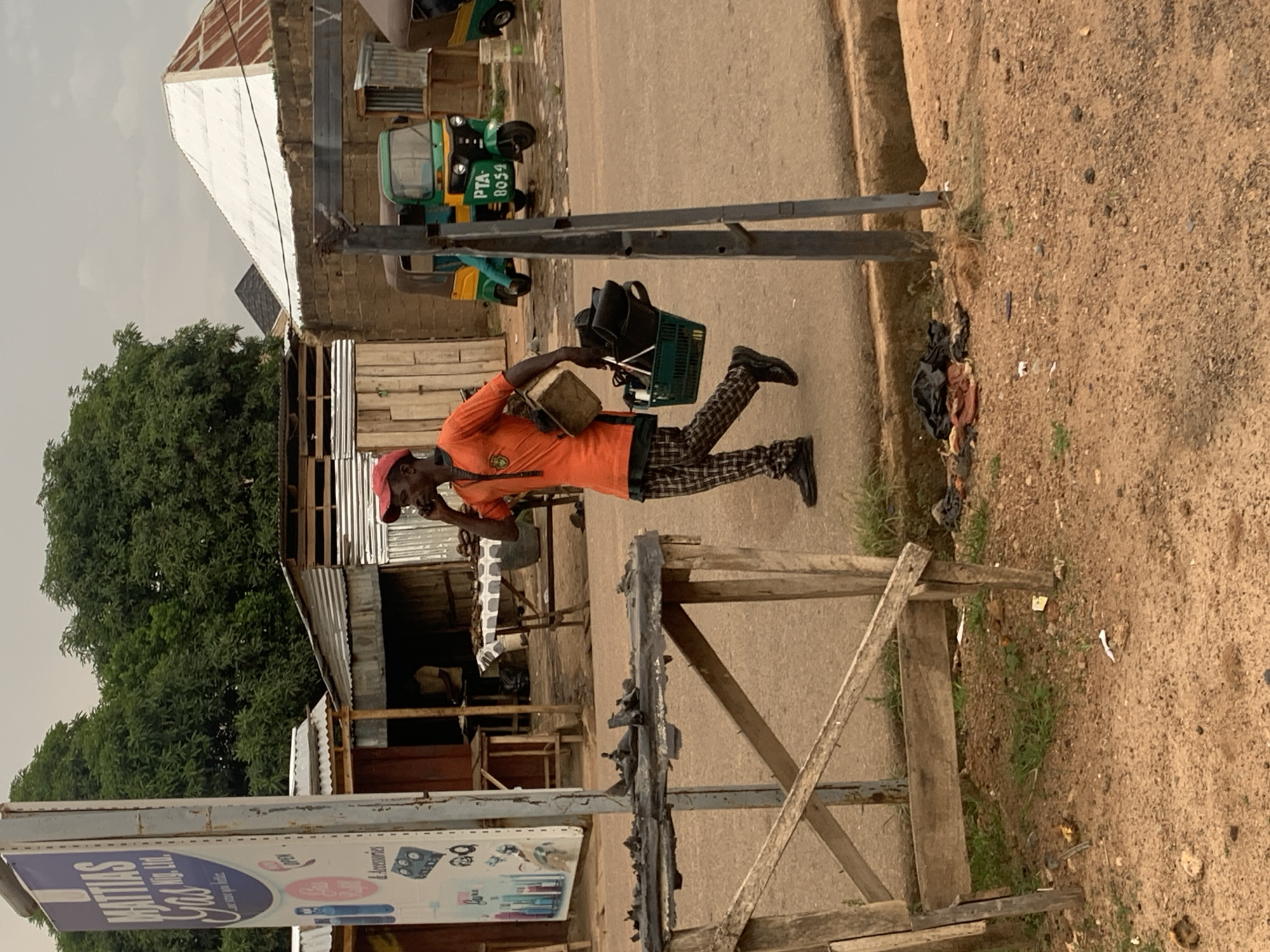 The image shows a man walking on a street in Jos, Nigeria. He is wearing an orange shirt, patterned pants, black shoes, and a red cap. He's carrying a box in one hand and holding a phone to his ear with the other. Behind him, there are several tuk-tuks (three-wheeled vehicles) parked on the side of the road. The area is dusty, with unpaved sections alongside the paved road. There are buildings of various construction types visible in the background, including some with corrugated metal. A sign for "Mattias Gas" is also visible. There's some garbage on the side of the road as well.
