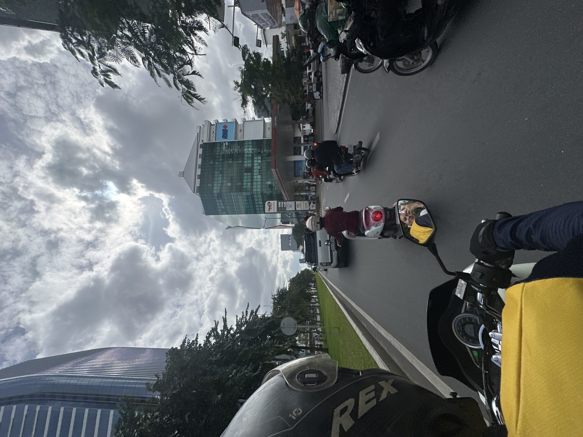 The image shows a street scene in Khan Daun Penh, Cambodia, captured from the perspective of someone riding a motorbike. In the foreground, a rider's black helmet, partially visible on the left, has the word "REX" printed on it. The rider's hands, wearing black gloves, are on the handlebars. A bright yellow bag is visible on the rider's lap. The road ahead is populated with several motorbikes and a white pickup truck.
In the middle ground, a motorbike with a rider wearing a red top is moving forward. To the right, another motorbike carries two individuals. Further ahead, a gas station with a red roof is visible, with several vehicles, including cars and motorbikes, present. Behind the gas station, a modern, multi-story building with a green glass facade stands tall. Another taller, blue-tinted skyscraper is visible on the far left. Trees line the side of the road, with a grassy median separating the lanes.
The sky is filled with dramatic, grey clouds, suggesting overcast weather. The overall scene depicts urban traffic in a city environment with modern architecture.