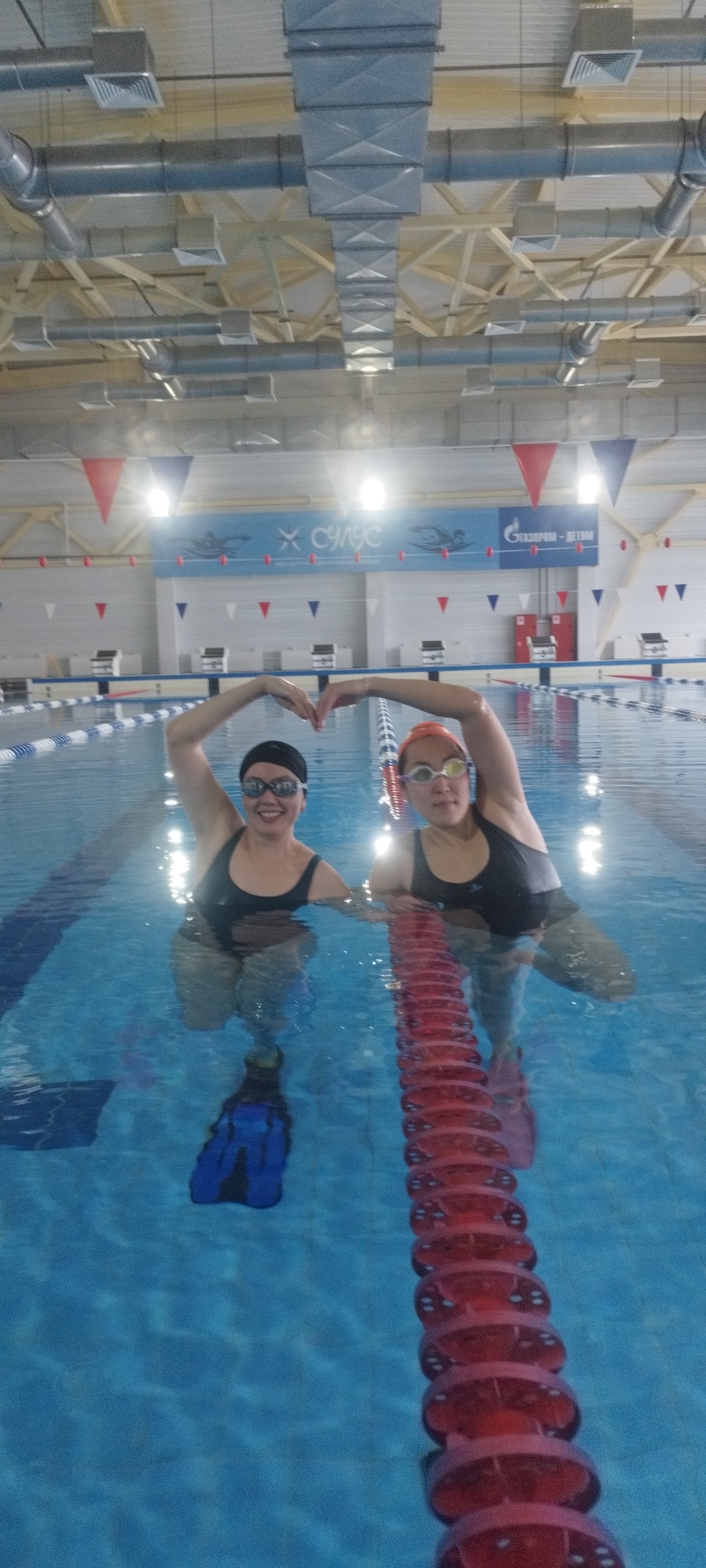 Two women in swimsuits and swim caps stand waist-deep in a swimming pool, forming a heart shape with their arms above their heads. The woman on the left wears black swim fins, a black cap, and swim goggles. The woman on the right wears an orange cap and swim goggles. The pool has lane dividers and blue and white tiles. The pool is at the Dolphin sports complex in Yakutsk, Russia. A banner with the names of sponsors is along the wall. The ceiling is high and contains metal ventilation ducts.