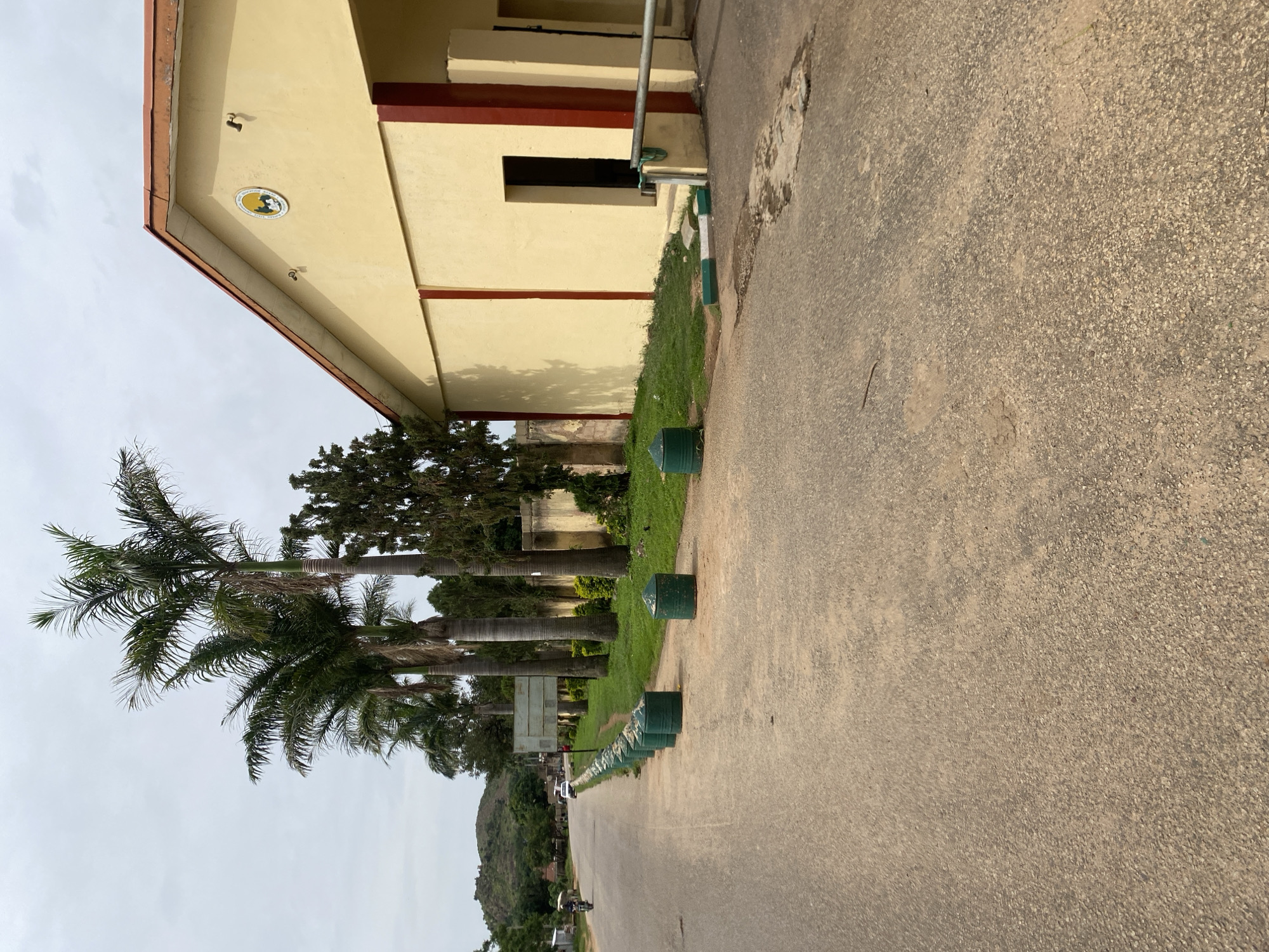 The image depicts a street scene in Jos, Nigeria, on an overcast day. The foreground is dominated by an asphalt road with some patches of wear and tear. Along the right side of the road, a line of green, cylindrical bollards with conical tops are spaced at intervals, bordering a grassy area. Behind the bollards is a low wall and the side of a building. The building is painted a pale yellow with dark red vertical accents on its corners and around an open doorway. A circular emblem with a silhouette of animals and trees is mounted on the building's facade. To the left of the road, several tall palm trees stand out against the sky, accompanied by other, darker green foliage and trees. In the distance, a motorcycle rider is visible on the road, and a white vehicle is parked further down. Hills with sparse vegetation can be seen in the far background. The overall impression is of a semi-urban or institutional setting.