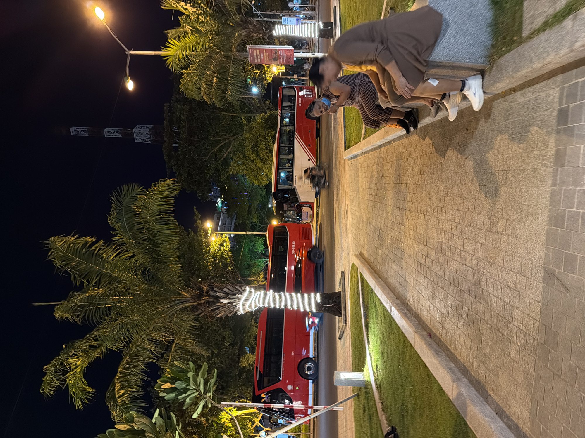 The image depicts a nighttime scene on a paved walkway in Nha Trang Ward, Vietnam. A group of people are seated on a concrete ledge. In the background, several red and white buses are parked on a street. Palm trees are visible throughout the scene, some of which are decorated with white string lights. A tall communication tower is also visible in the distance. The walkway is illuminated by streetlights, casting shadows across the pavement.