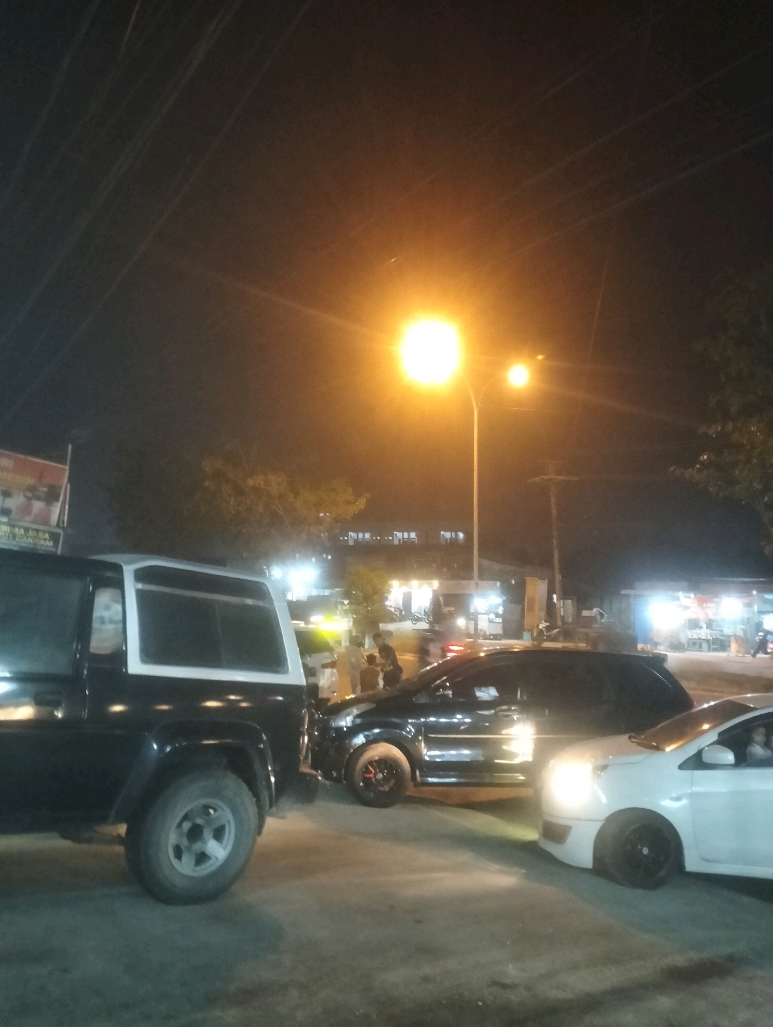 In Duri Barat, Indonesia, at night, a dark-colored SUV is parked on the left, facing right. To its right, a black car is parked, also facing right, with its rear bumper close to the SUV's front. A white car is parked to the right of the black car, facing right, with its front end nearly touching the black car's rear. The scene is illuminated by streetlights and lights from nearby businesses. A street sign is visible on the left, and trees are present in the background. Several individuals are standing or walking around the vehicles and businesses.