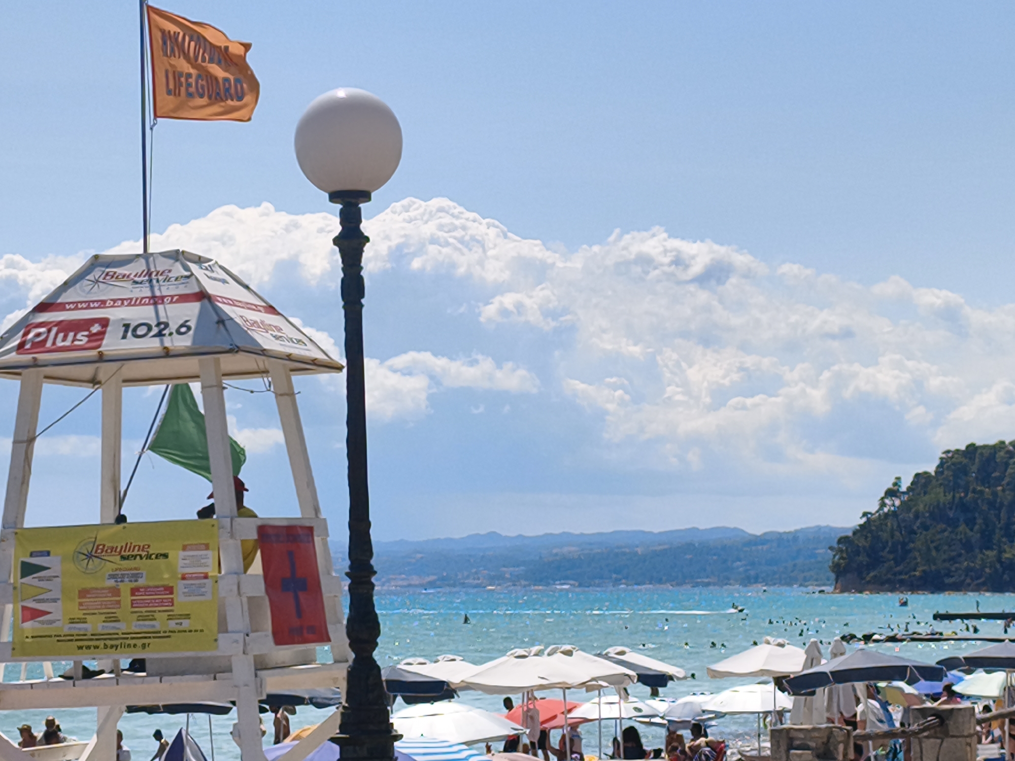 A bright, sunny day at a crowded beach in Kassandra Municipal Unit, Greece. The foreground is dominated by a white lifeguard tower adorned with signs and flags. A prominent orange flag with the word "LIFEGUARD" in black lettering flutters above the tower. Below, a yellow sign with "Bayline services" and various symbols is displayed, along with a red flag featuring a white cross. A lamppost with a spherical light fixture stands to the right of the tower.
The beach itself is filled with numerous beach umbrellas, providing shade to people lounging on the sand. The turquoise sea stretches out behind the beach, dotted with swimmers and boats, including a speed boat in the distance. In the background, lush green hills and mountains rise, creating a picturesque coastal landscape under a blue sky with scattered white clouds. The scene captures the lively atmosphere of a popular summer beach destination.
