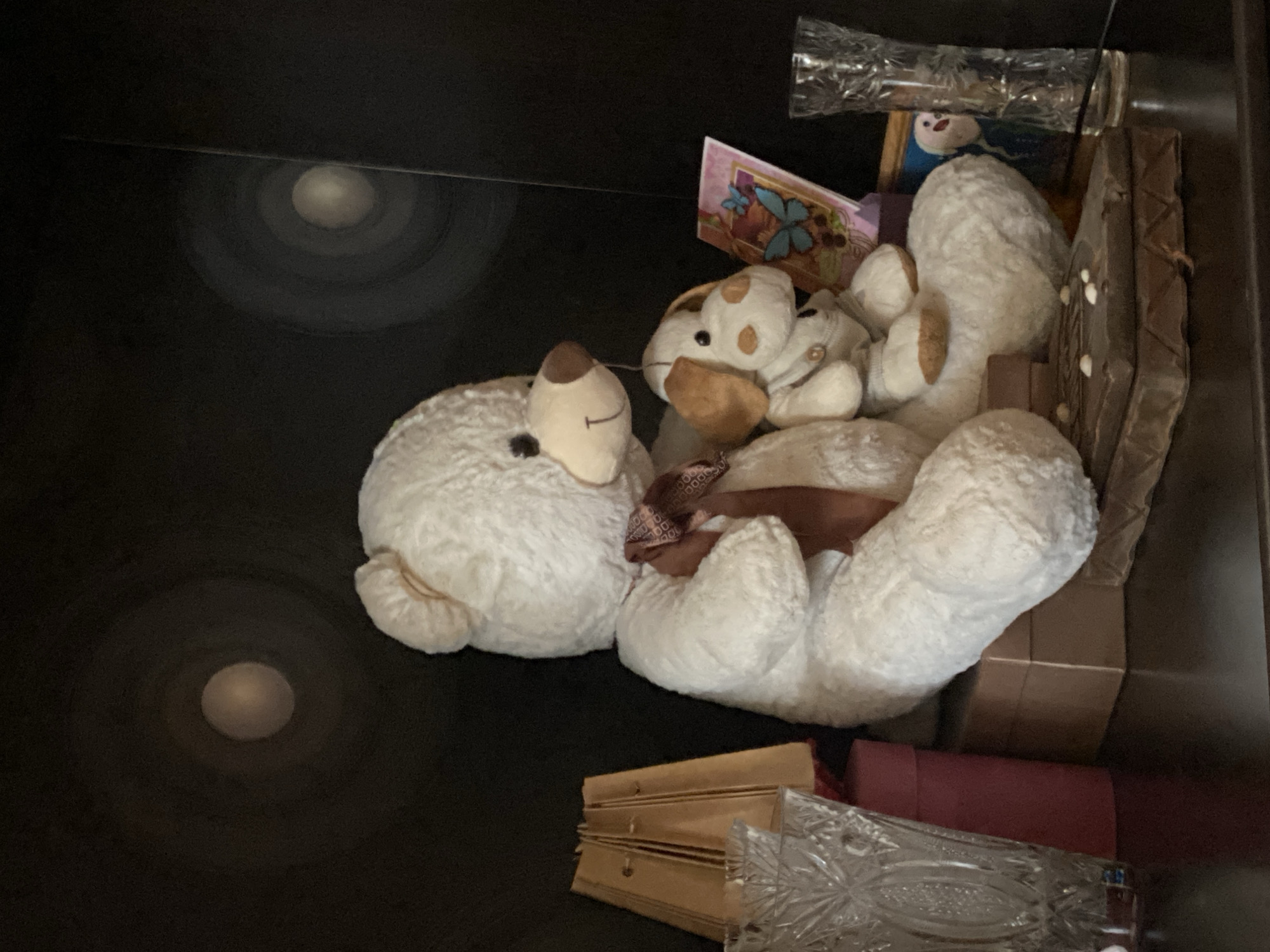 This media file shows a collection of stuffed animals and other objects stored in what appears to be a dark drawer or box. The main subjects are two large, white, fluffy stuffed animals, likely sheep or similar creatures, positioned in the center of the frame. One is lying on its side, with its head resting on its paws, while the other is partially visible behind it. Nestled between them is a smaller, light-colored stuffed dog with brown spots. A pink greeting card with a floral and butterfly design is propped up against the stuffed animals, with a small snowman figurine visible behind it.
Other objects in the scene include a dark brown, chocolate bar-shaped object with white details on the right, a stack of gold-colored folders or boxes in the lower right, a red cylindrical object, and a clear, patterned glass object that appears to be a vase or decanter. A shiny, crinkled material, possibly plastic wrap, is also visible at the top right. The background is dark and nondescript. Two circular light reflections are present on the dark surface on the left side of the image, suggesting a light source above and to the left, or a glossy surface reflecting light.
No activity or event is taking place; the objects appear to be stored. There are no visible people. The lighting suggests an indoor setting, likely with artificial light. The time of day and weather are not discernible. Location cues are limited to the provided context of Novaya Usman', Russia, which is not visually evident in the image itself. No text is clearly legible. The overall mood is one of storage and perhaps a sense of childhood nostalgia due to the presence of stuffed animals.