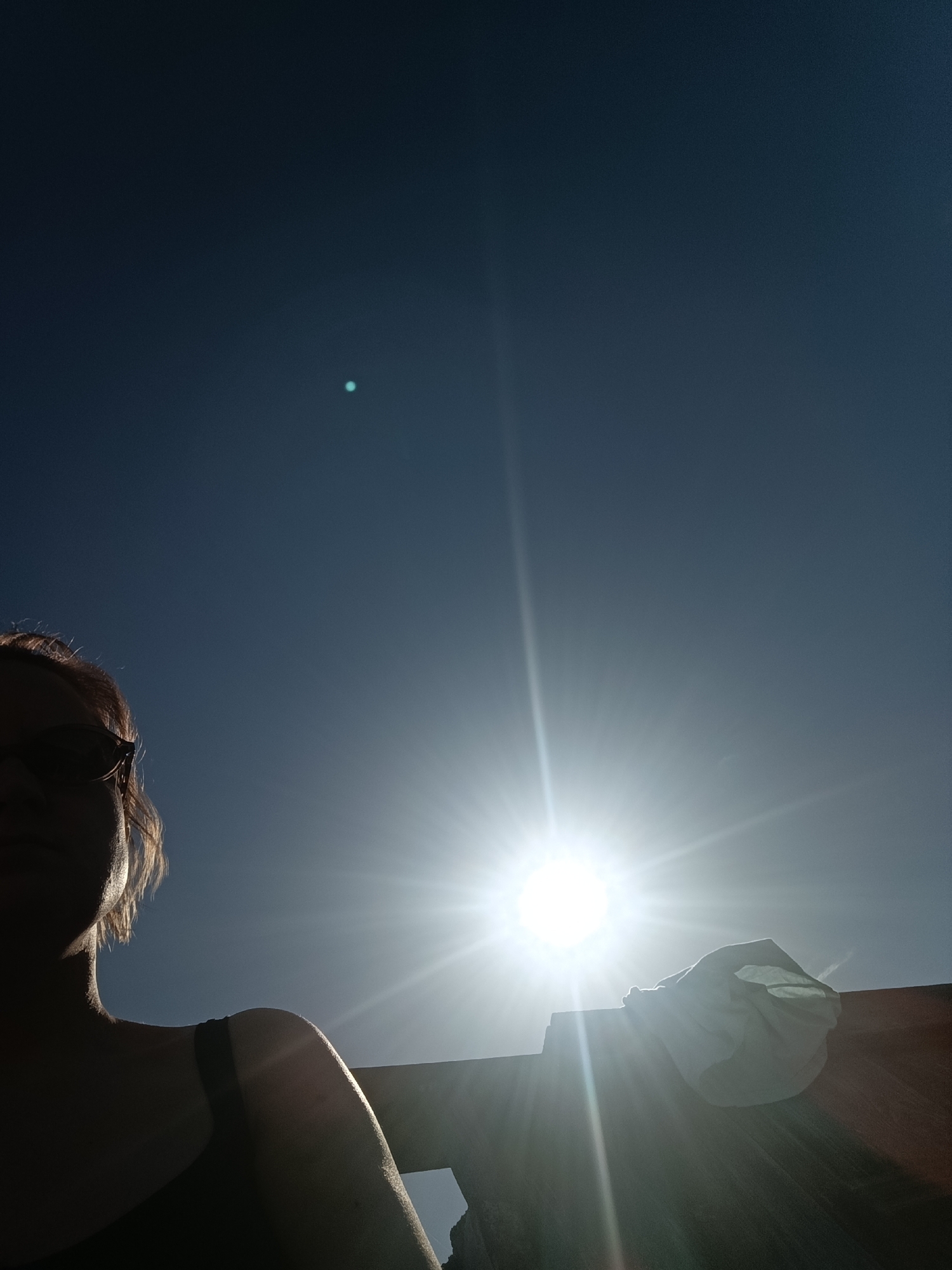 A low-angle shot captures a person in silhouette against a bright, sunlit sky. The person, presumably a woman, is wearing sunglasses and her shoulder is bare. To the right, a white cloth or garment is draped over what appears to be a wooden fence or railing. The sun is positioned centrally in the frame, emitting intense rays. The sky is a clear dark blue with a small, distant bright dot above the sun. The location is unknown.